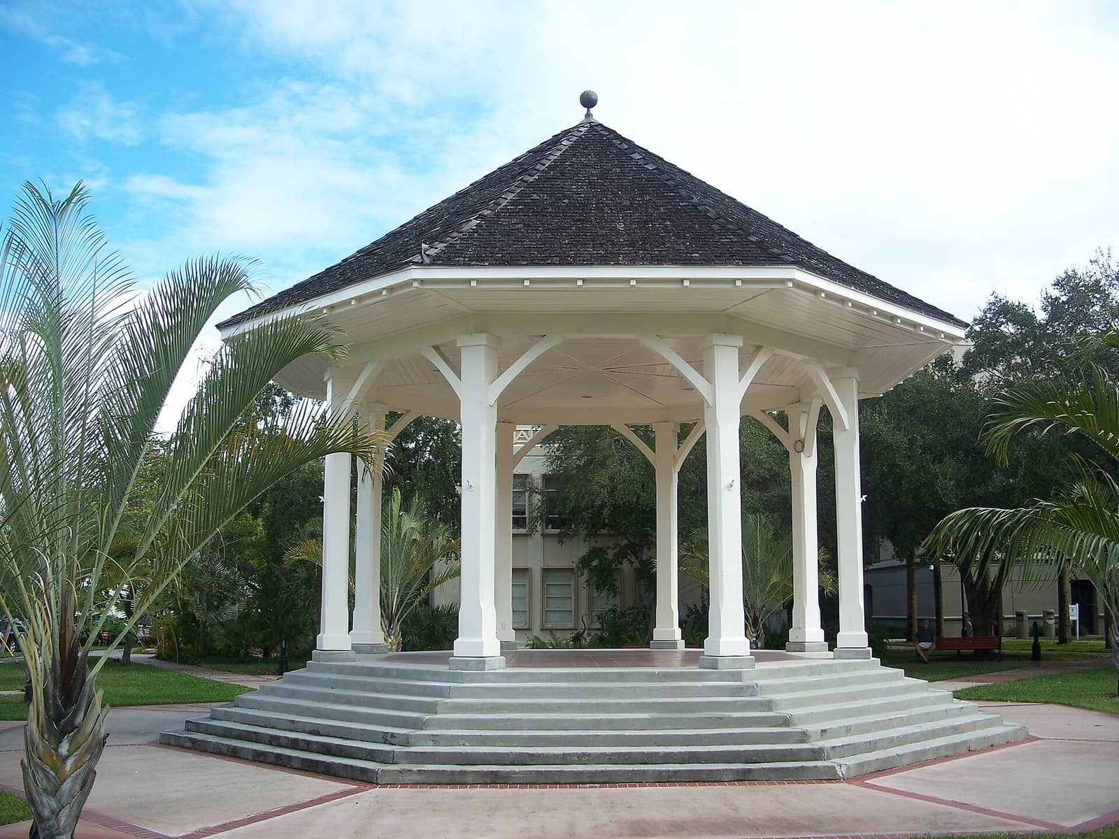White gazebo with dark shingled roof and stone steps under a blue sky with clouds.
