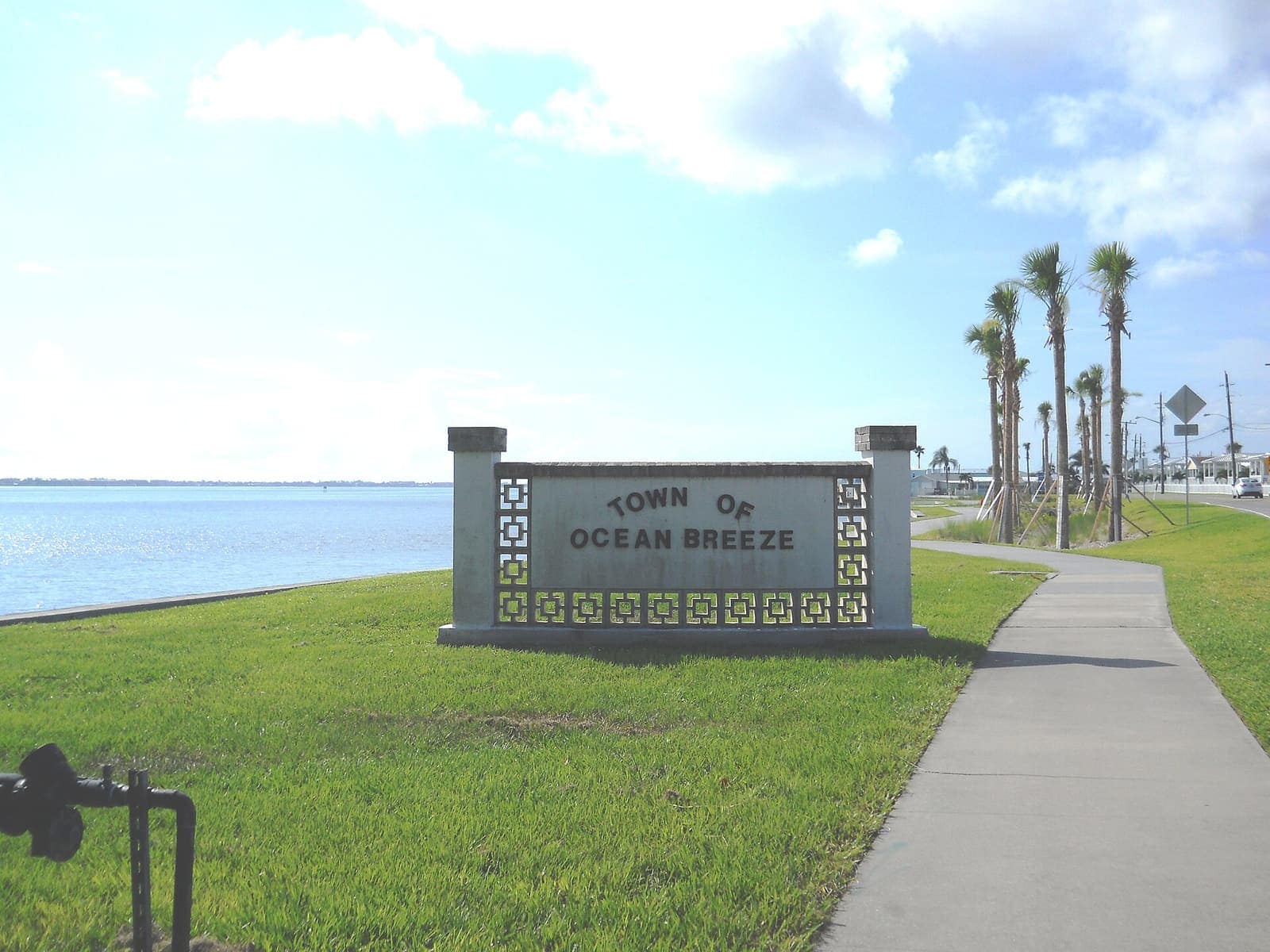 Concrete Town of Ocean Breeze welcome sign beside a grassy lawn, calm water, palm trees, and a sidewalk.