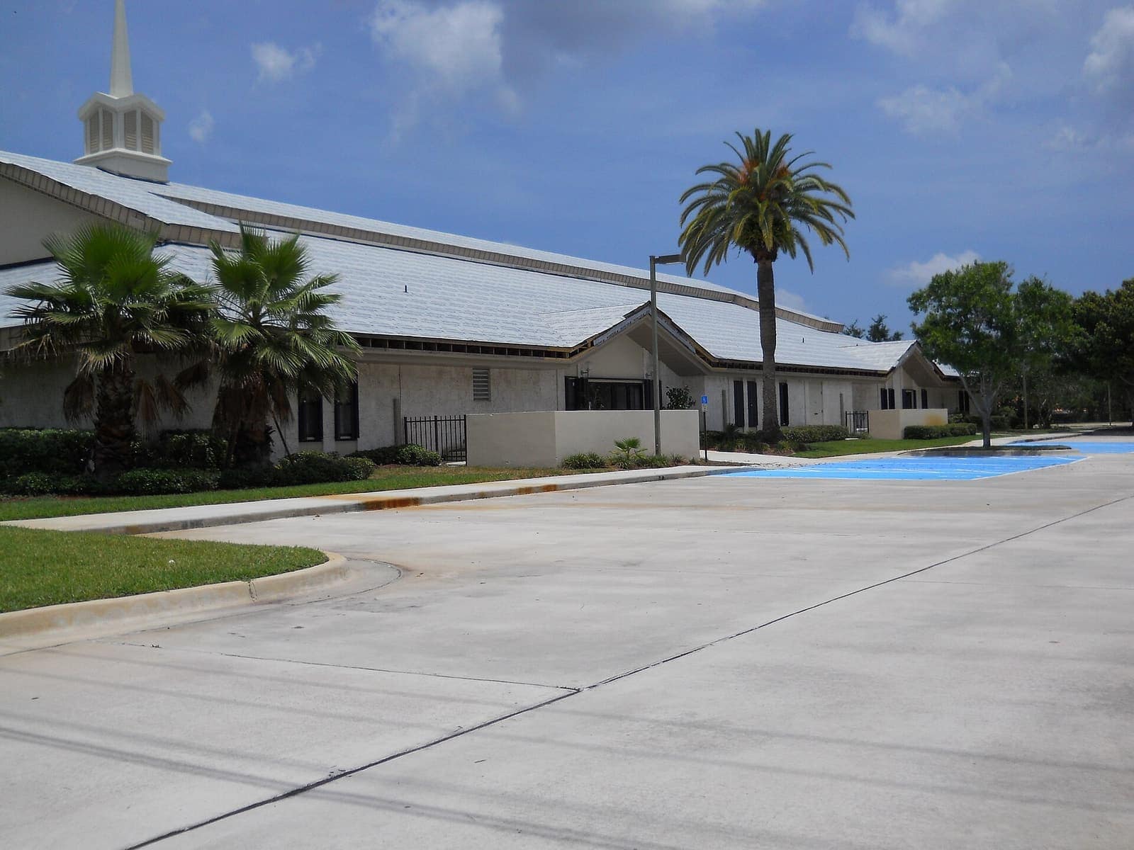 White single-story church building with a steeple, palm trees, and a wide concrete driveway in daylight.