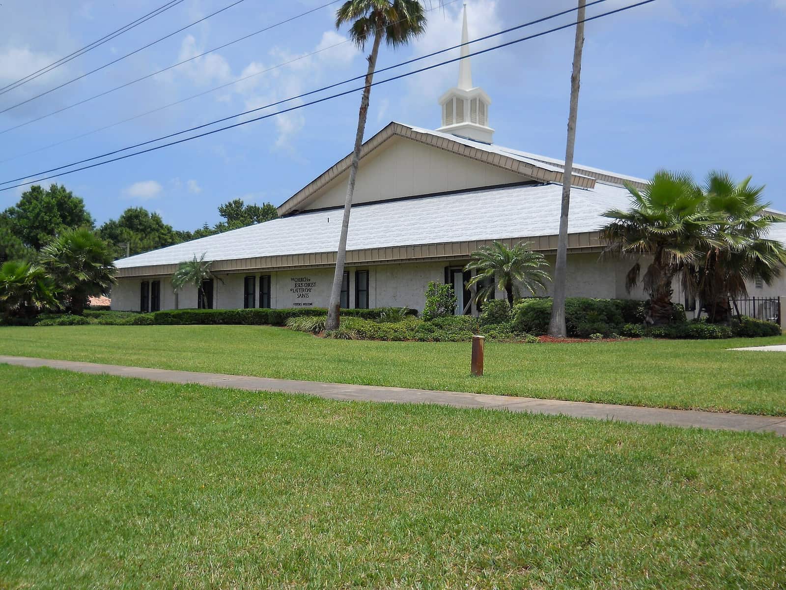 White church building with a steeple and metal roof beside a grassy lawn, palm trees, and power lines overhead.