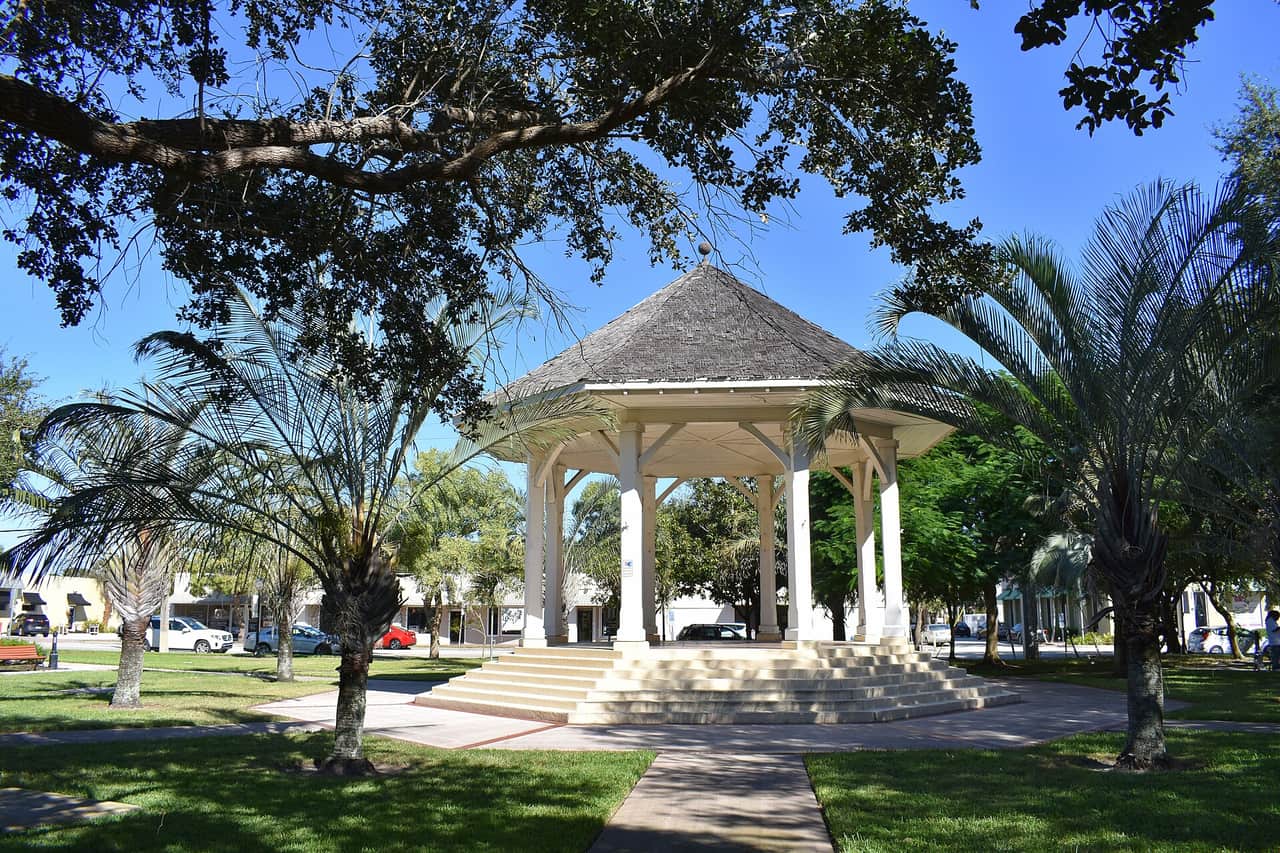 White domed gazebo with stone steps in a green park under palm trees and blue sky.