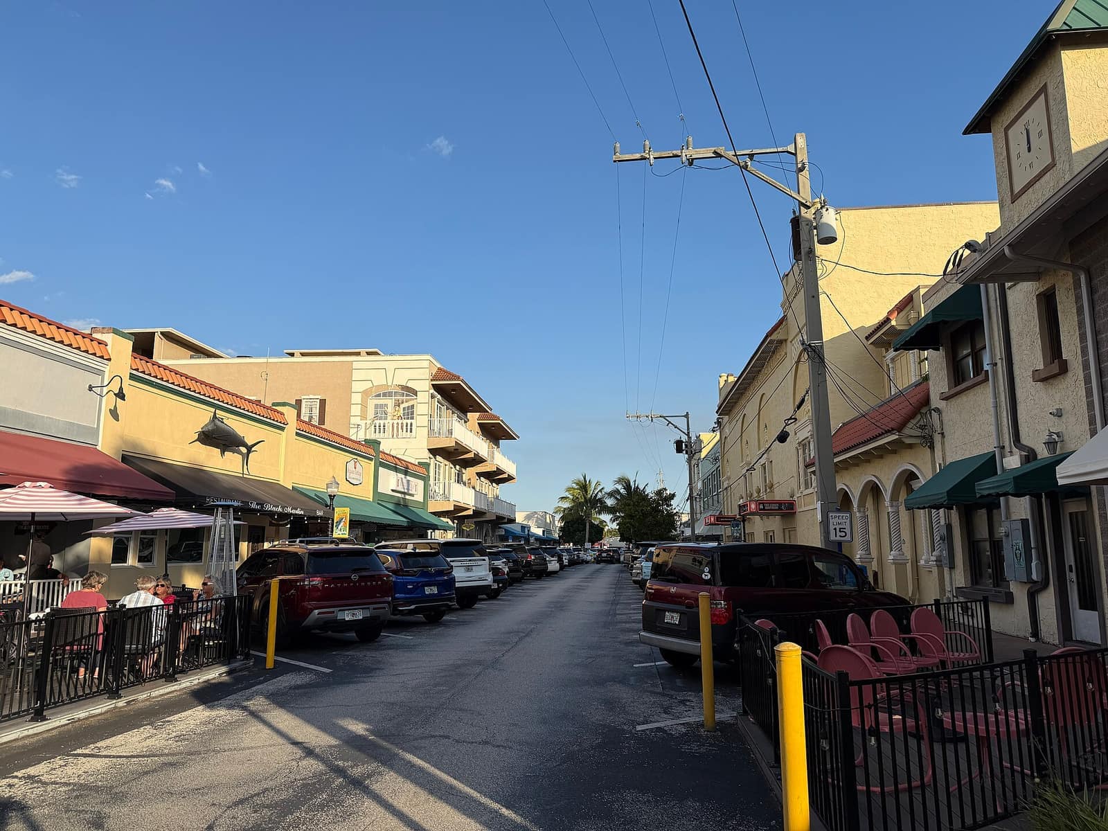 Downtown street with parked cars, storefronts and awnings under a clear blue sky in late afternoon light.