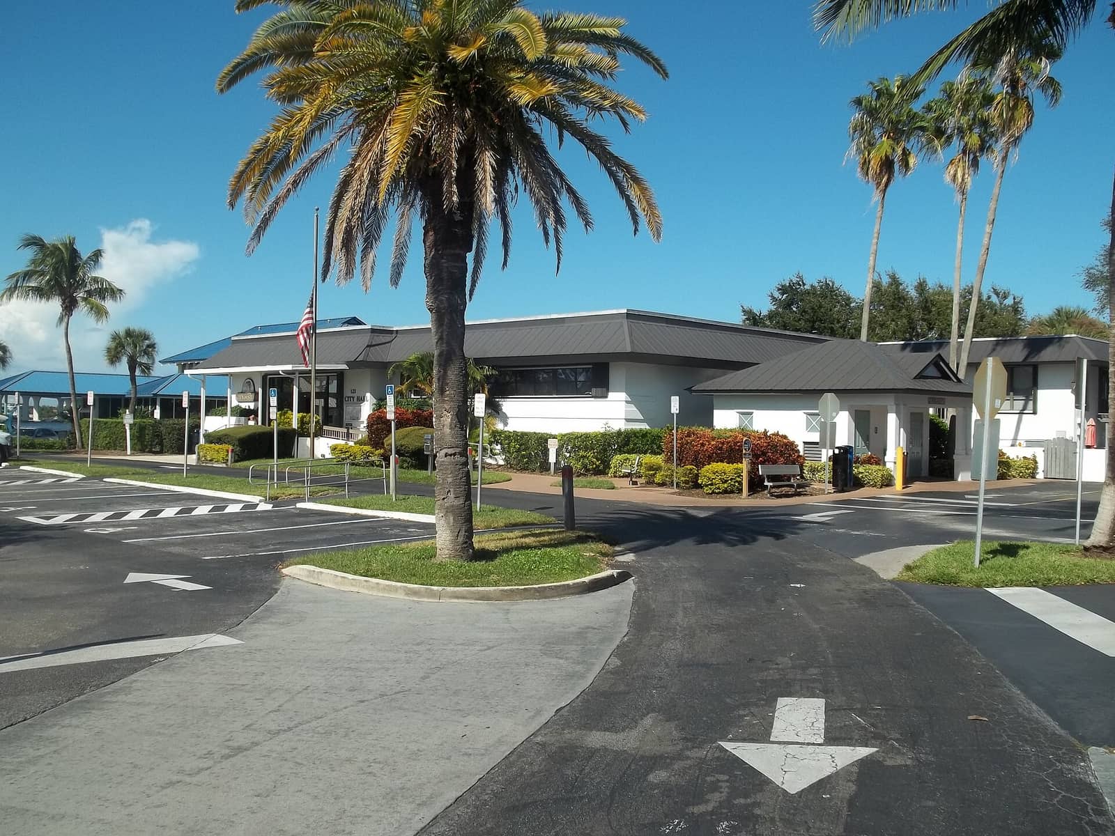 Single-story white municipal building with dark roof behind palm trees and a flagpole.