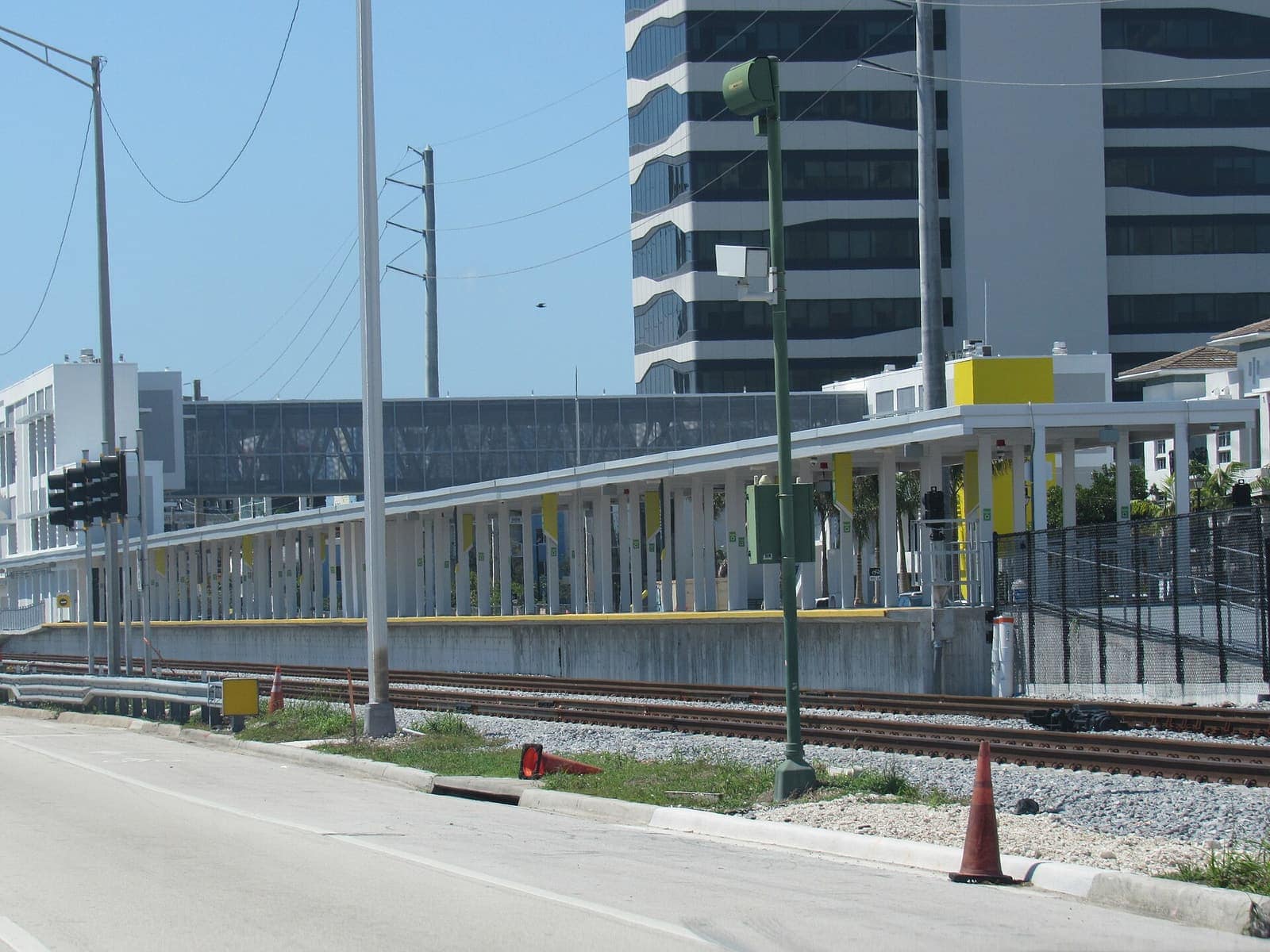 Elevated train platform with yellow-trimmed canopy beside tracks, with a high-rise tower behind.