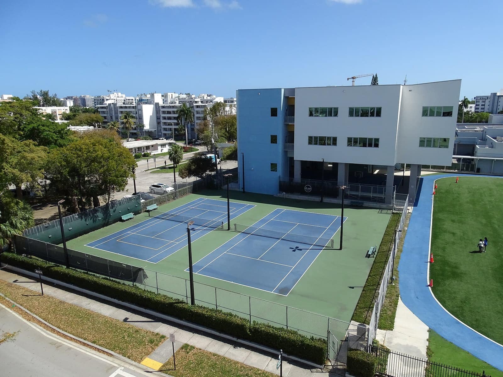Two blue tennis courts beside a blue and white building, viewed from above on a sunny day.