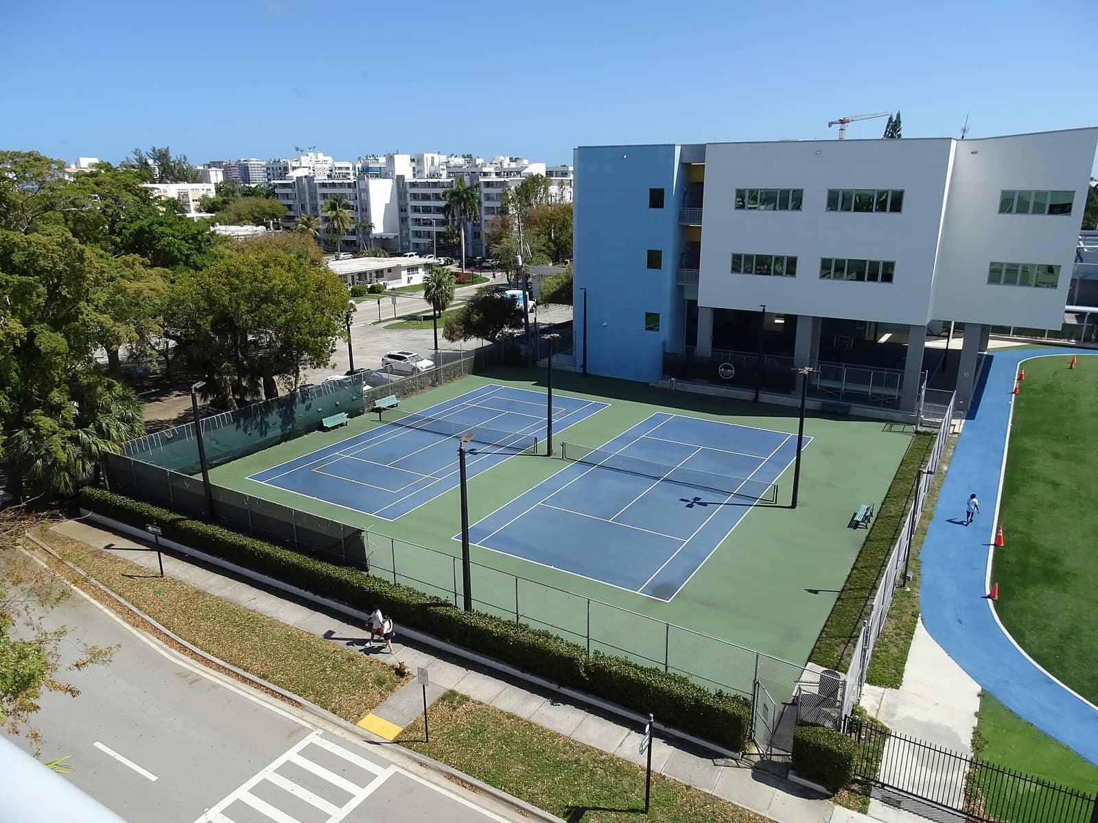 Overhead view of two tennis courts beside a modern blue and white building under clear skies.
