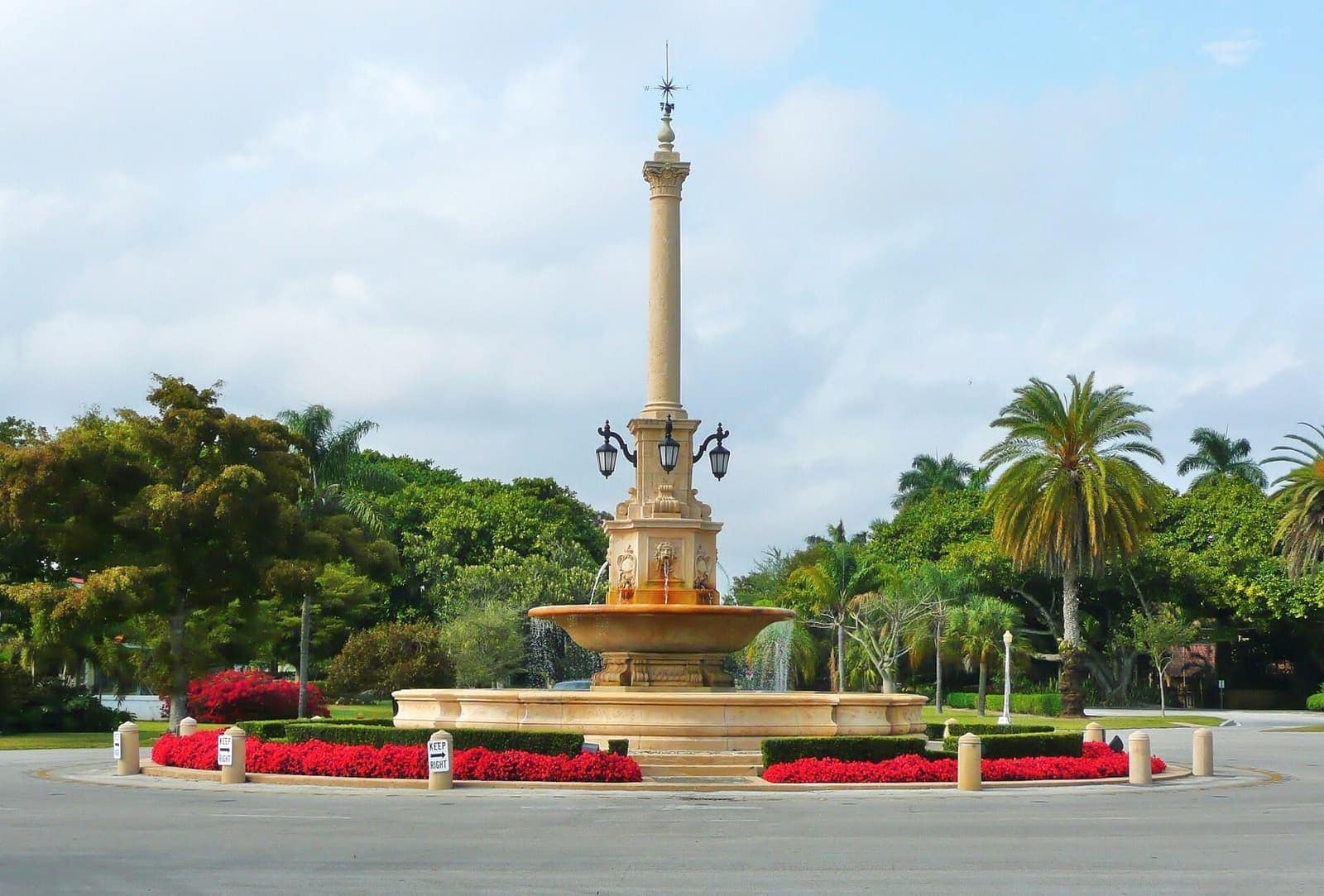 Tall stone column monument over a circular fountain, ringed by red flowers and palm trees under a cloudy sky.