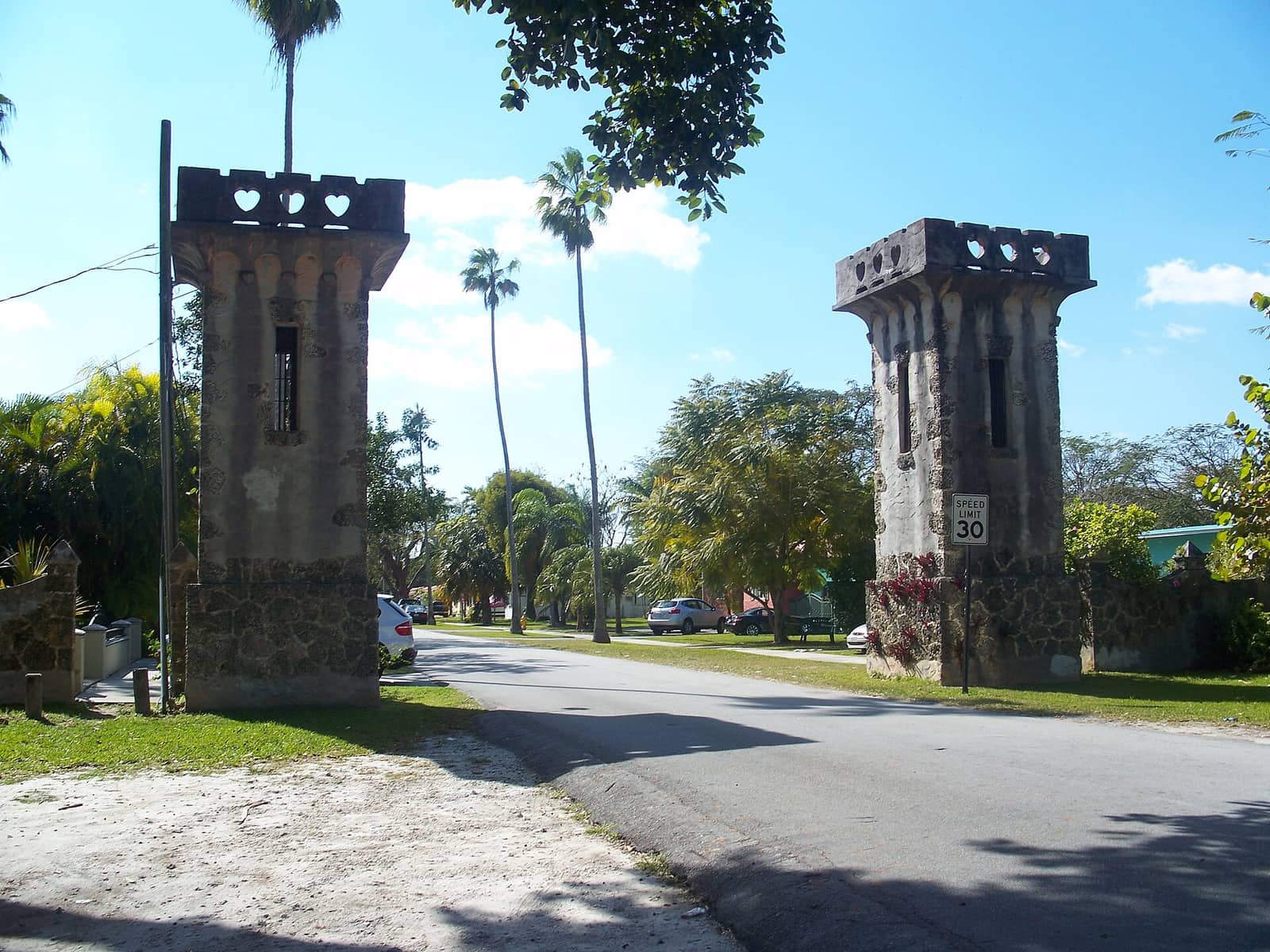 Two stone gate towers with heart-shaped cutouts flank a tree-lined road with parked cars.
