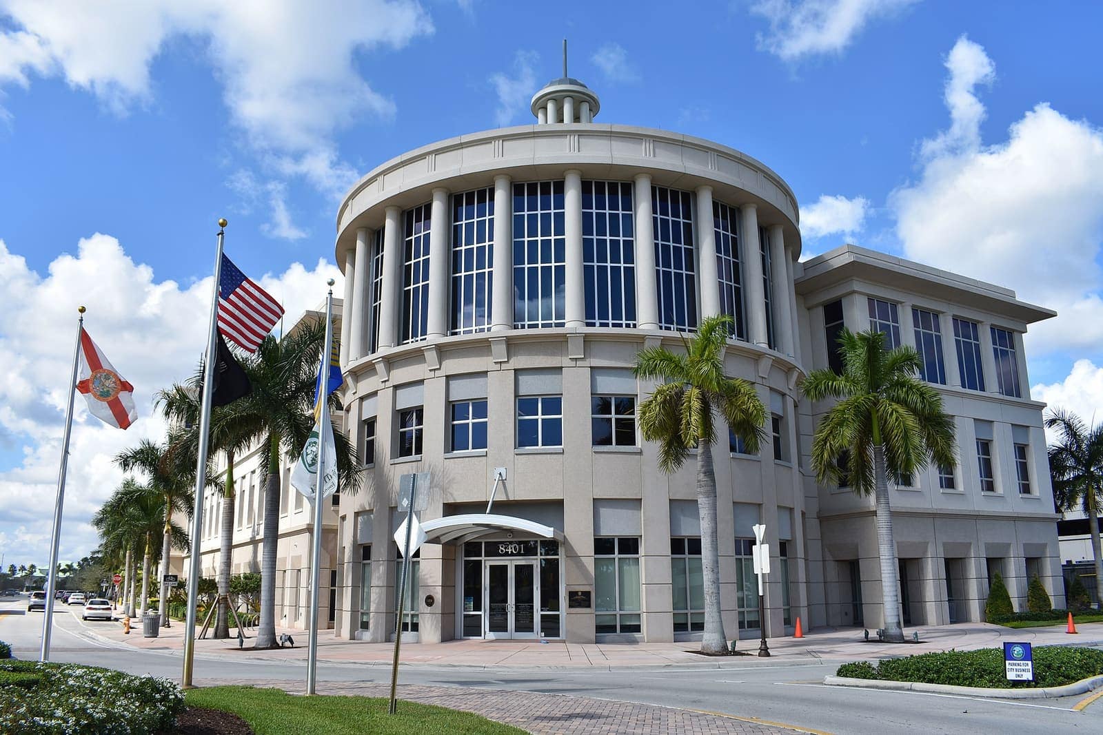 Curved cream-colored government building with glass windows, flags, and palm trees under a blue sky.