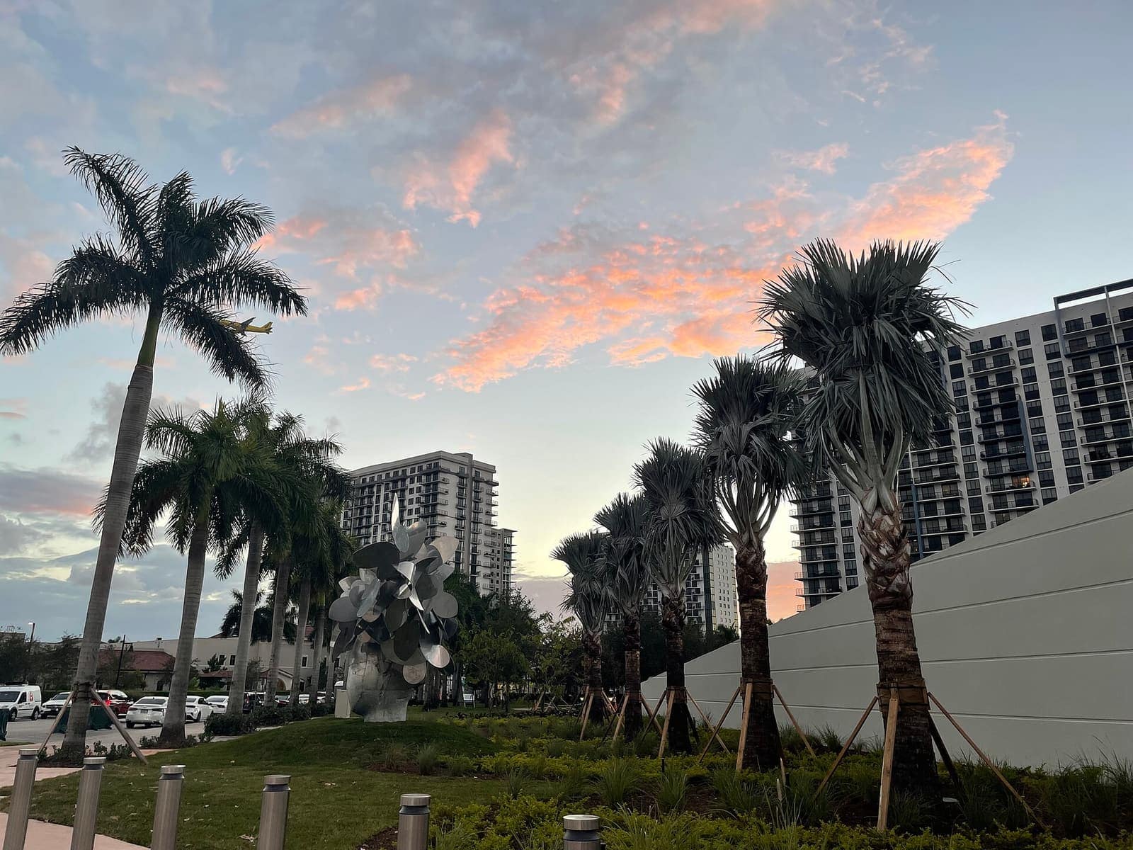 Pink-tinted evening sky above tall palms, a metal sculpture, and high-rise buildings along a walkway.