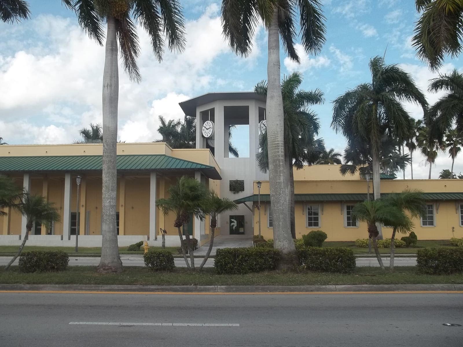 Yellow two-story city hall building with a clock tower, green roof trim and tall palm trees.