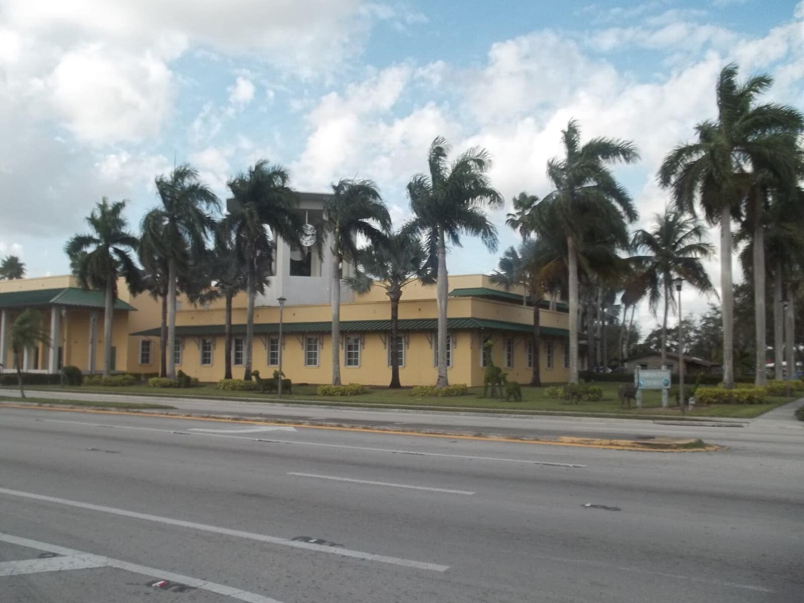 Yellow city hall building behind a row of palm trees along a wide daytime road.