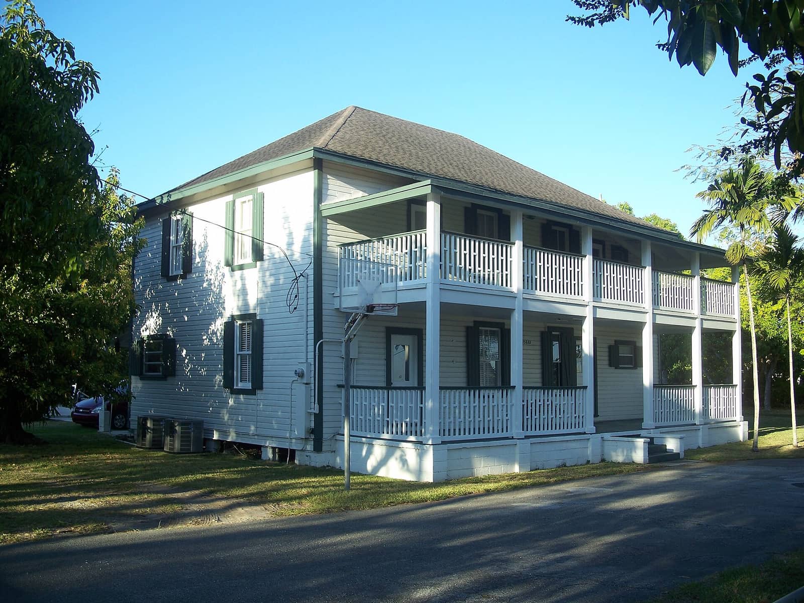 White two-story wooden house with long porches, shaded by trees under a clear blue sky.