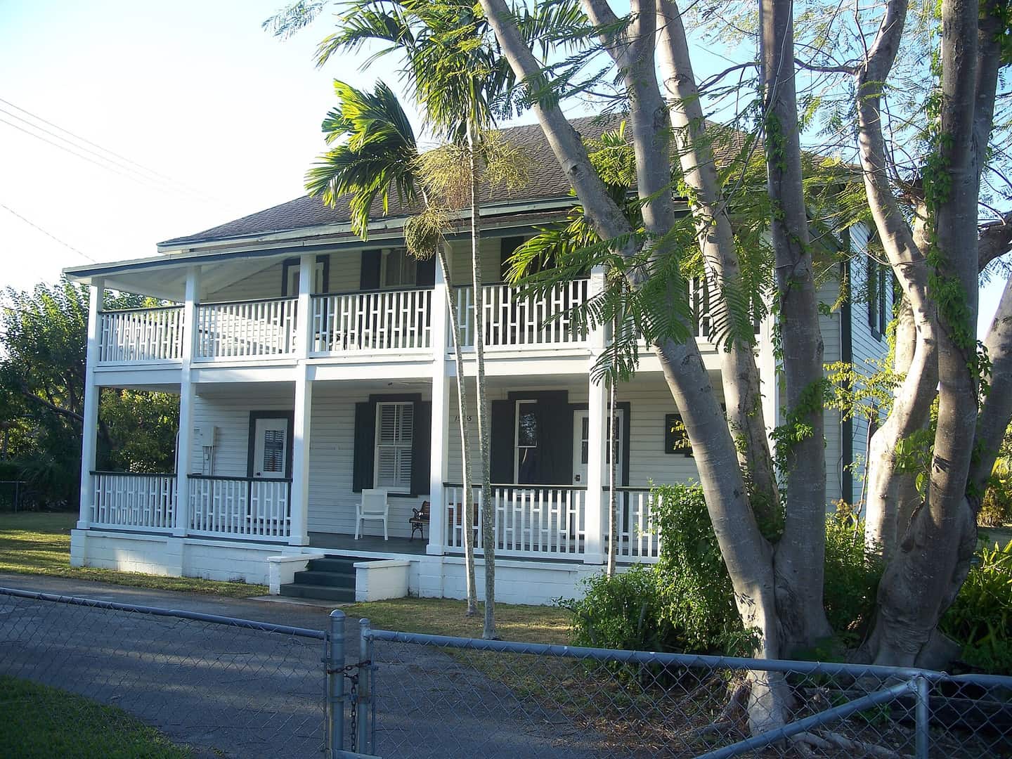 White two-story wooden house with columned porches, palm trees, and a chain-link fence in front.