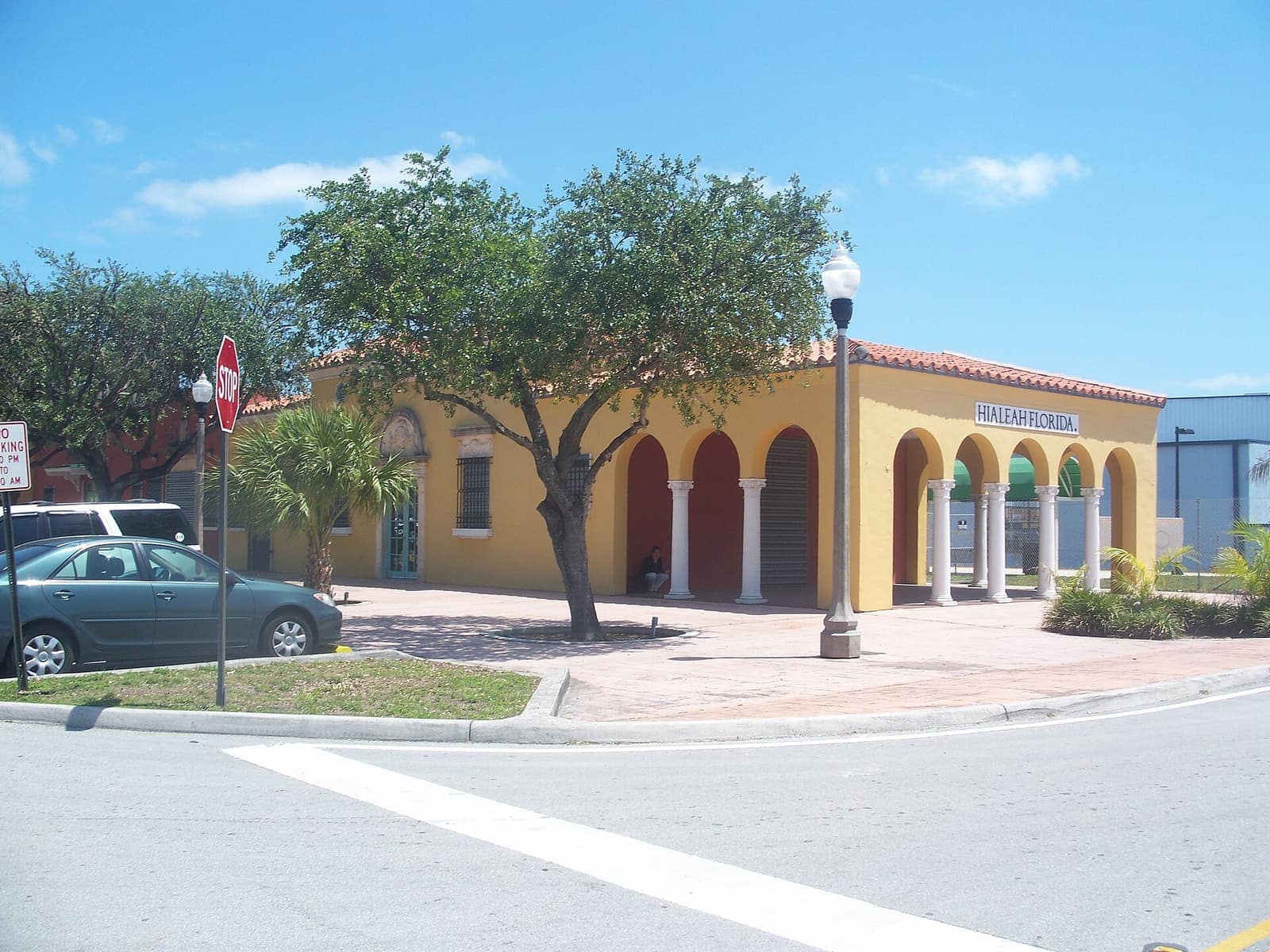 Yellow stucco former railway station with arched colonnade and red-tiled roof beside a large tree and street.