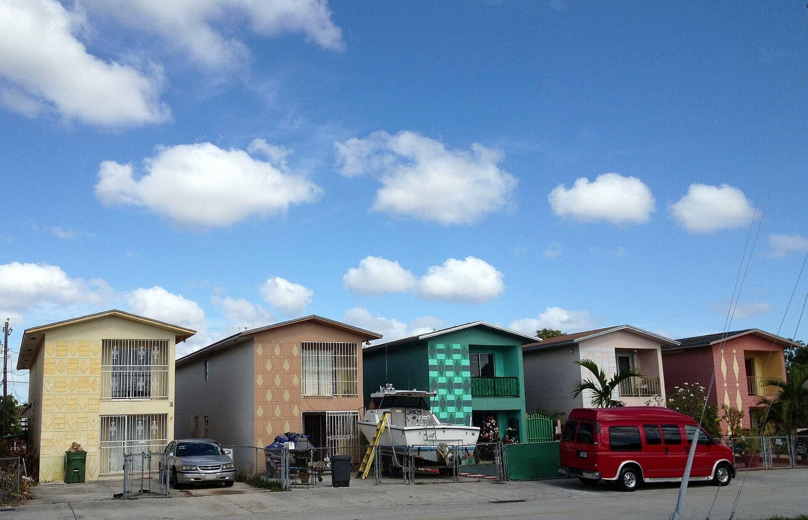 Row of colourful two-storey houses with barred windows, parked cars and a red van under blue sky with clouds.