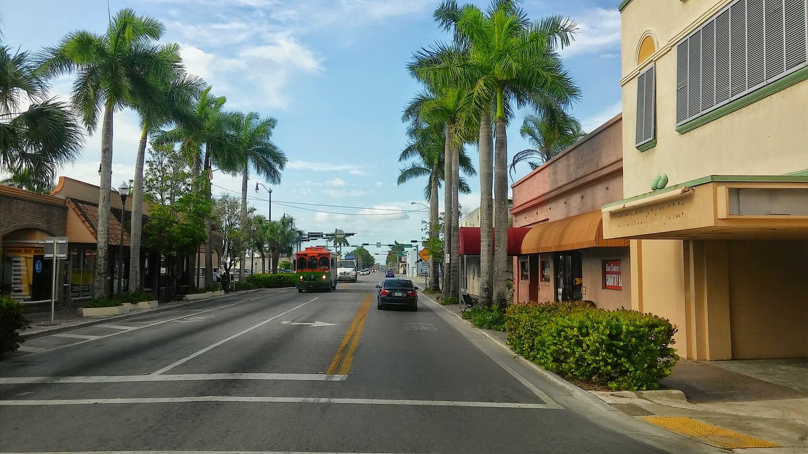 Palm-lined downtown street with low storefronts, a red truck and a car driving toward the distance.