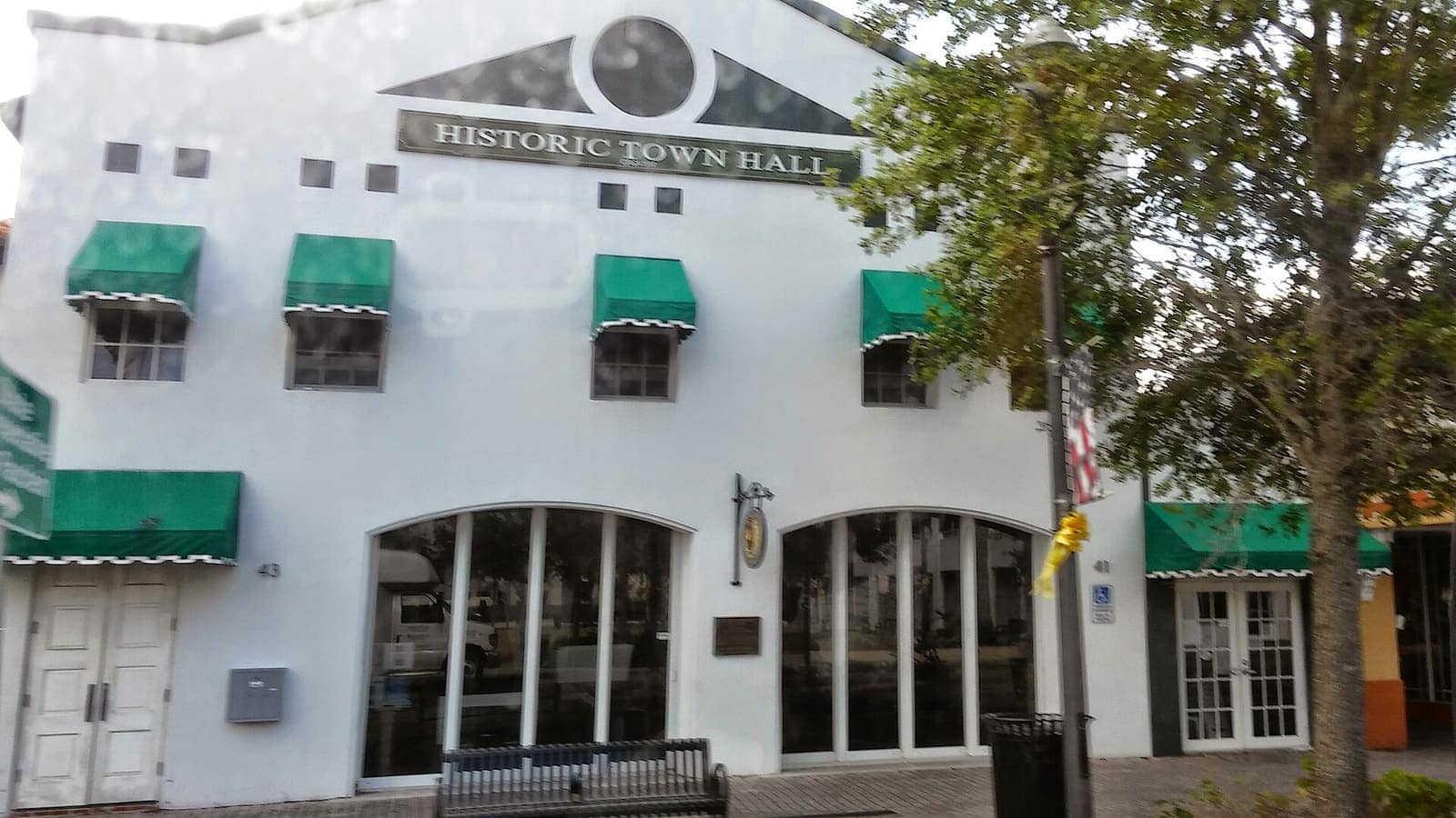 White stucco historic town hall with green striped awnings, arched windows and a bench on the sidewalk.