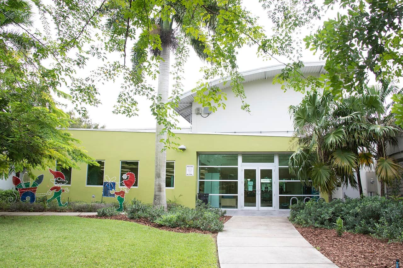 Glass entrance doors of a pale yellow building with painted figures and palm trees in front.