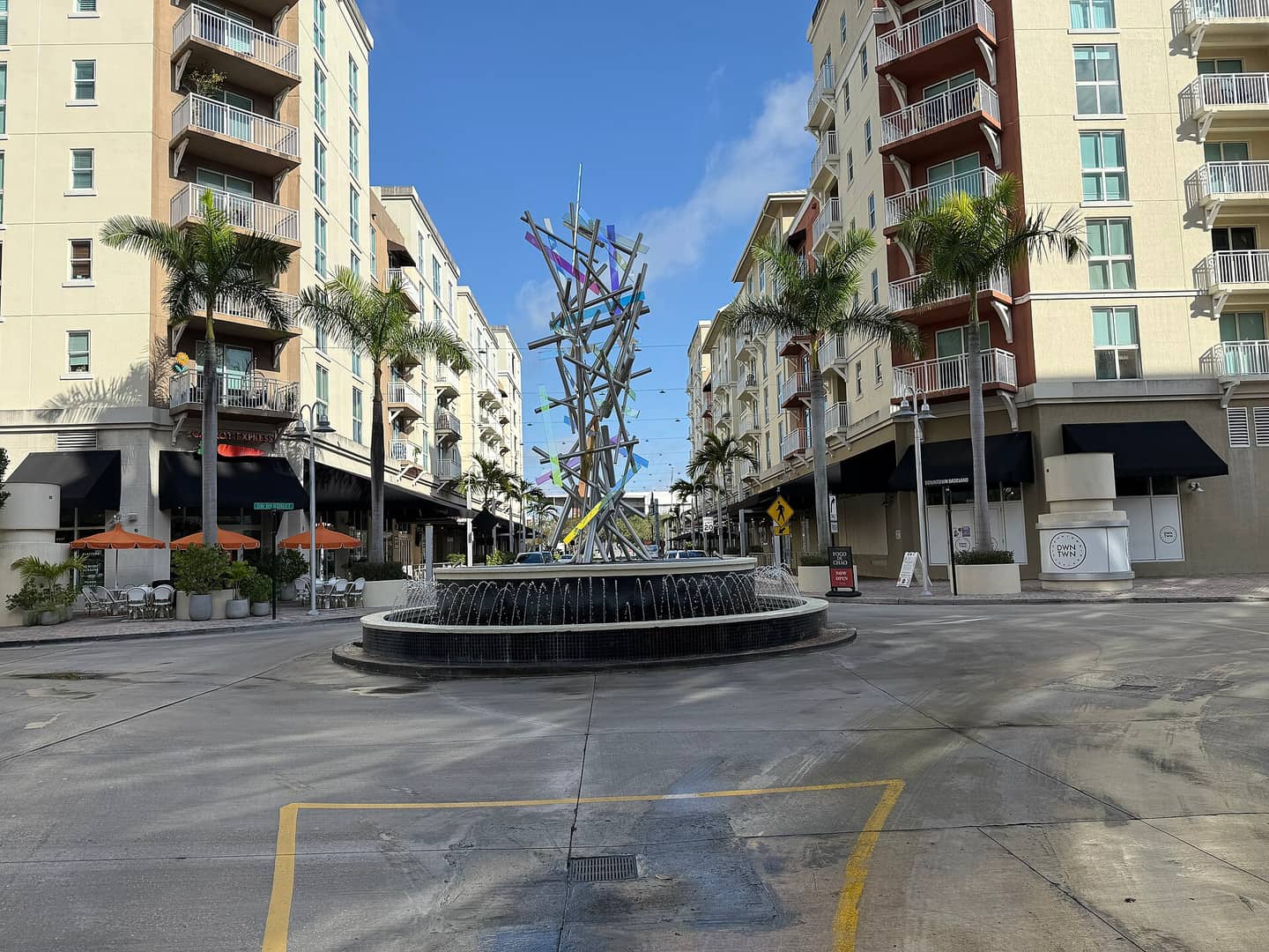 Roundabout with a decorative sculptural fountain surrounded by palm trees and mid-rise buildings.