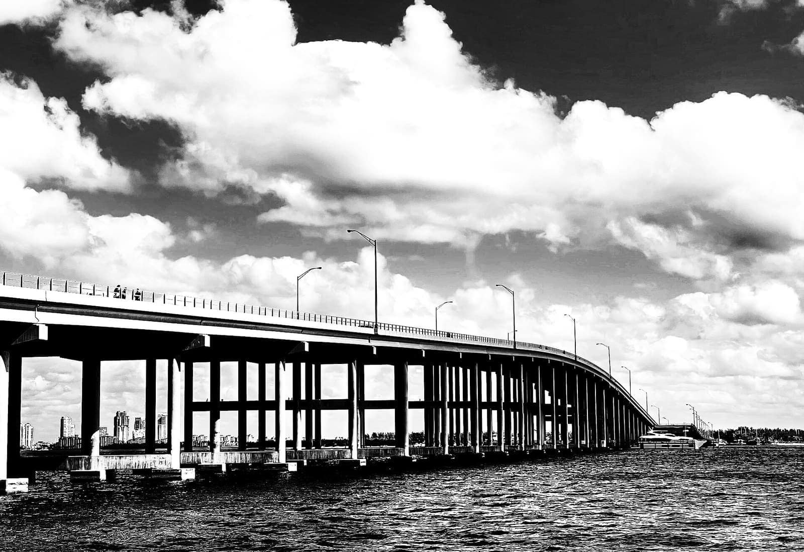 Black and white view of a long concrete causeway bridge over water under big clouds.