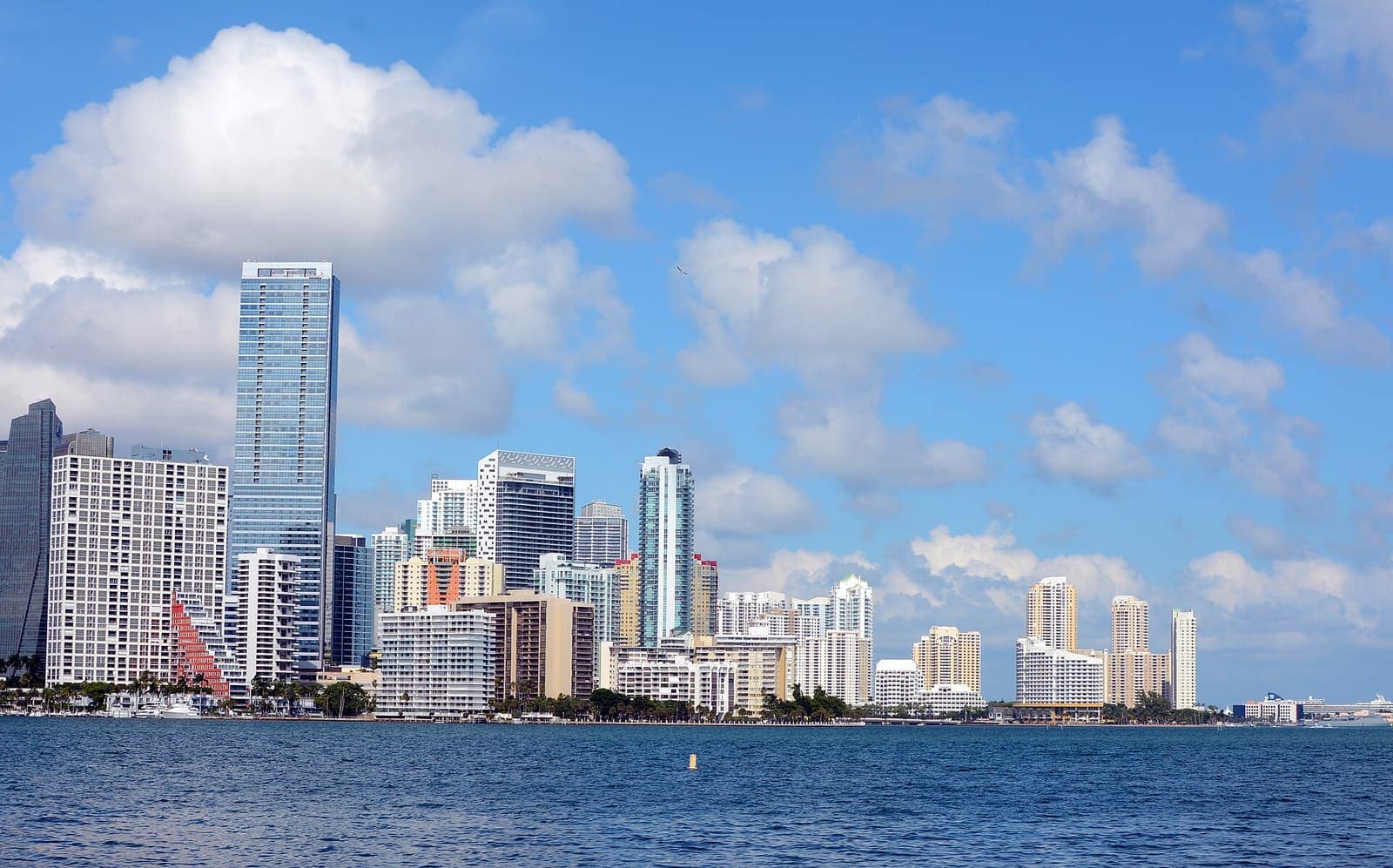 Downtown skyline of tall glass towers rising above blue water under a partly cloudy daytime sky.