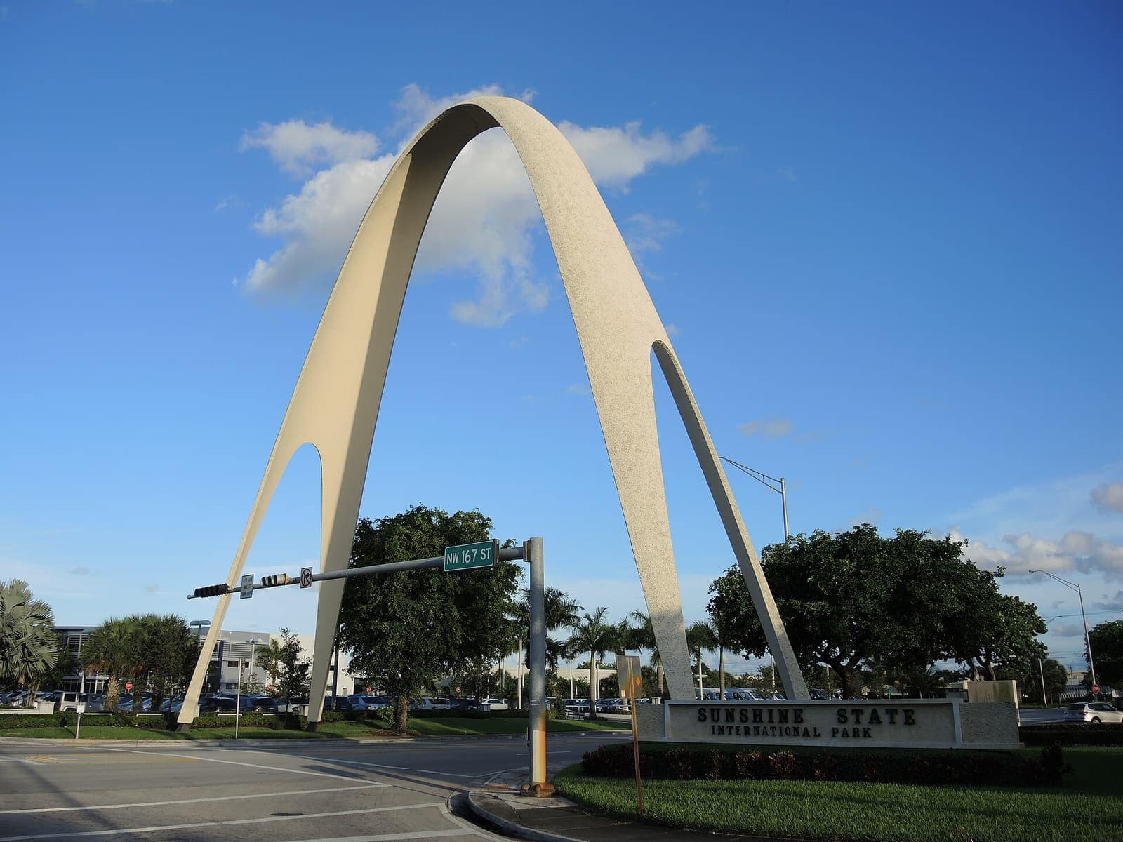 Tall pale concrete arch over an intersection, with a Sunshine State International Park sign and a blue sky.