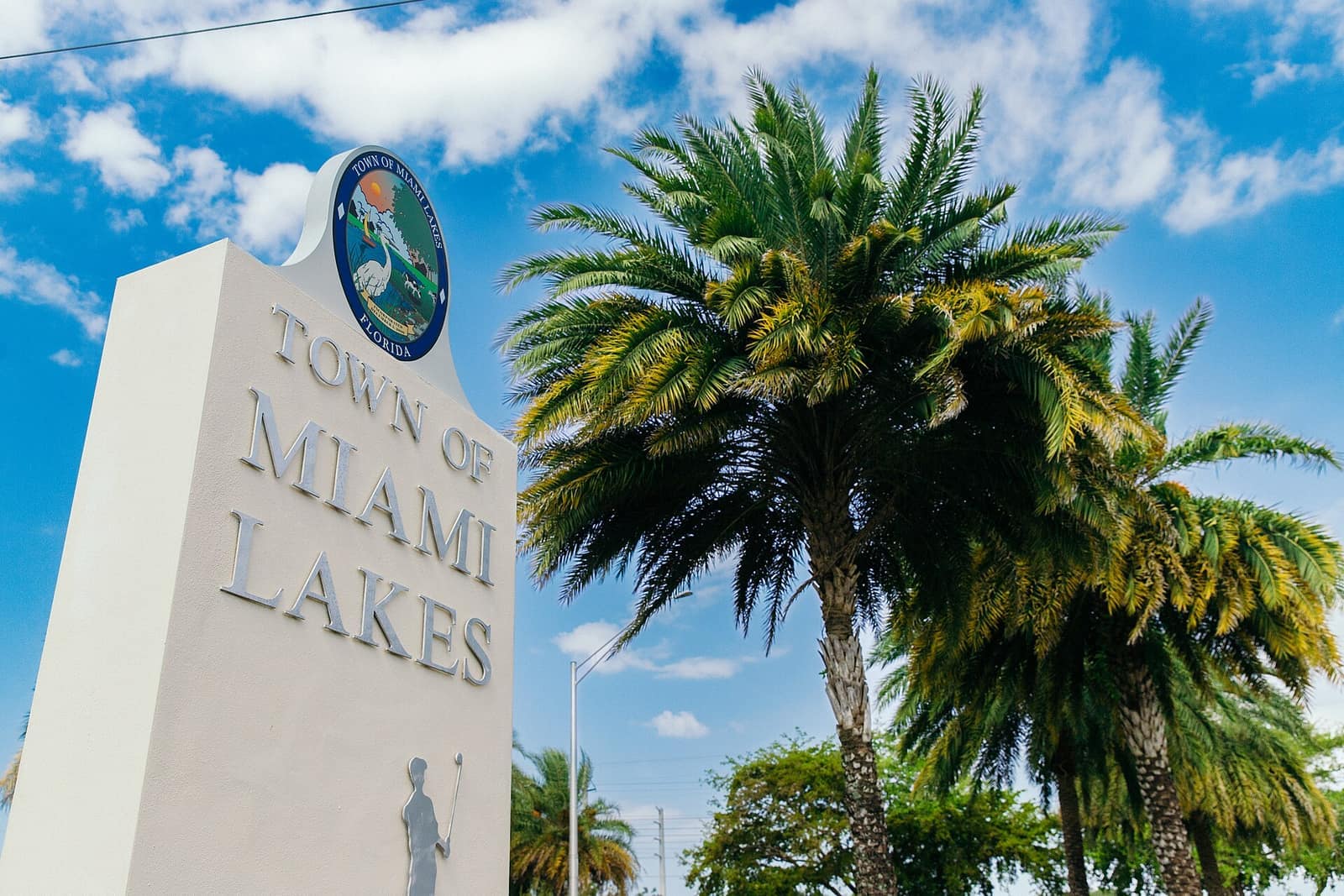 A white Town of Miami Lakes entrance sign stands beside tall palm trees under a blue sky.