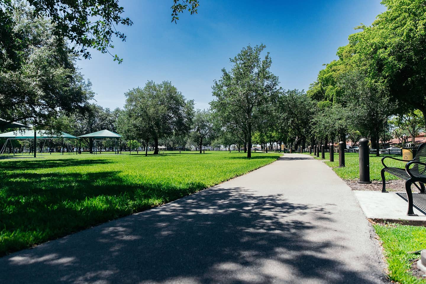 A paved walkway runs through a green park lawn lined with shade trees, benches, and pavilions.