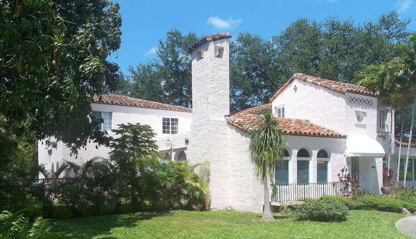 White stucco Mediterranean-style house with terracotta tile roof and chimney tower behind a green lawn.