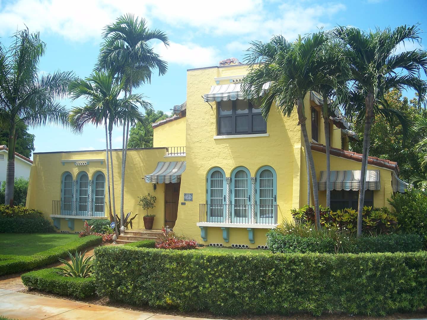 Yellow stucco house with arched blue-trimmed windows, tall palms and a clipped hedge in front.