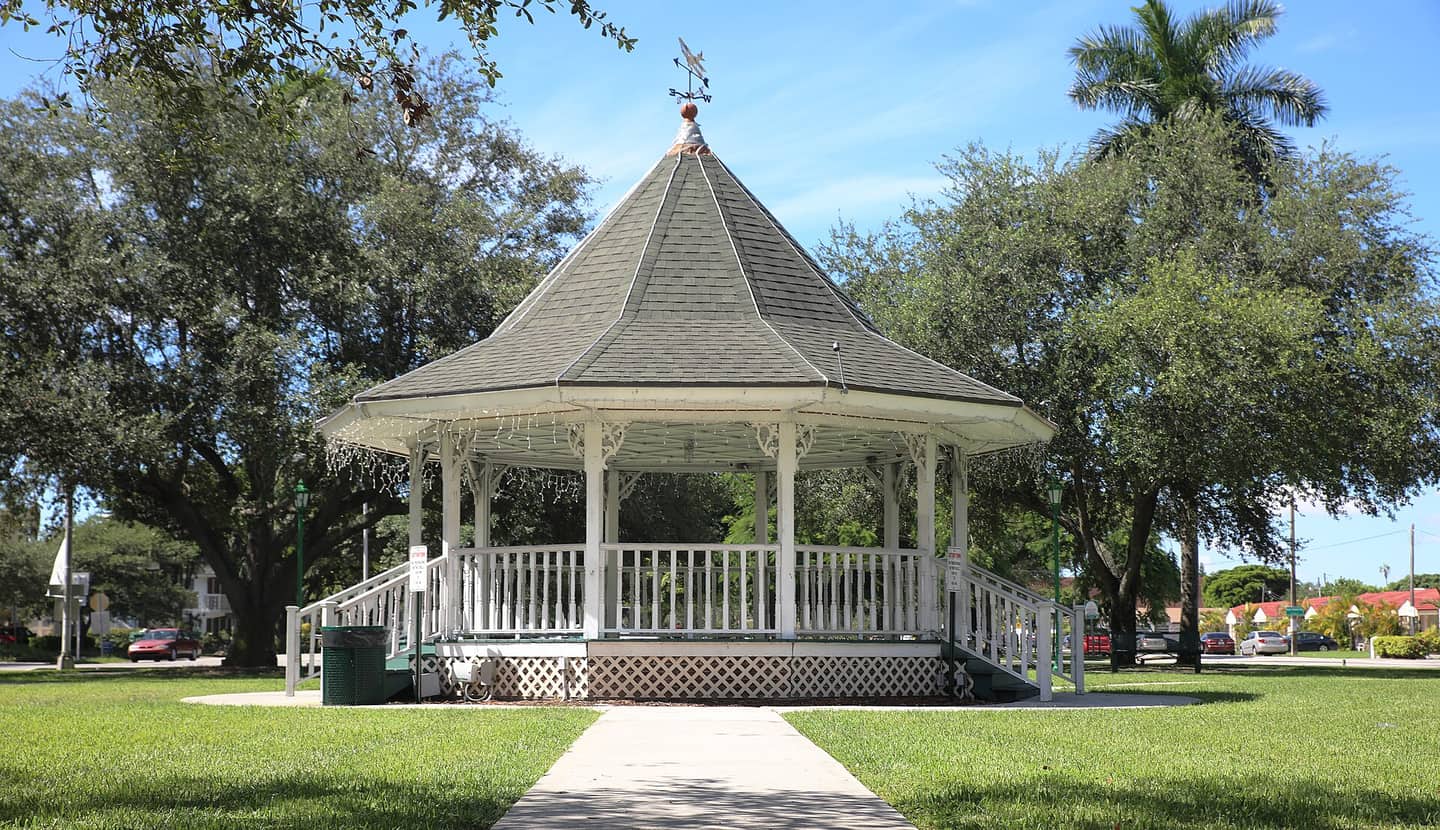 White wooden gazebo with a grey shingled roof and weathervane on green lawn under trees.