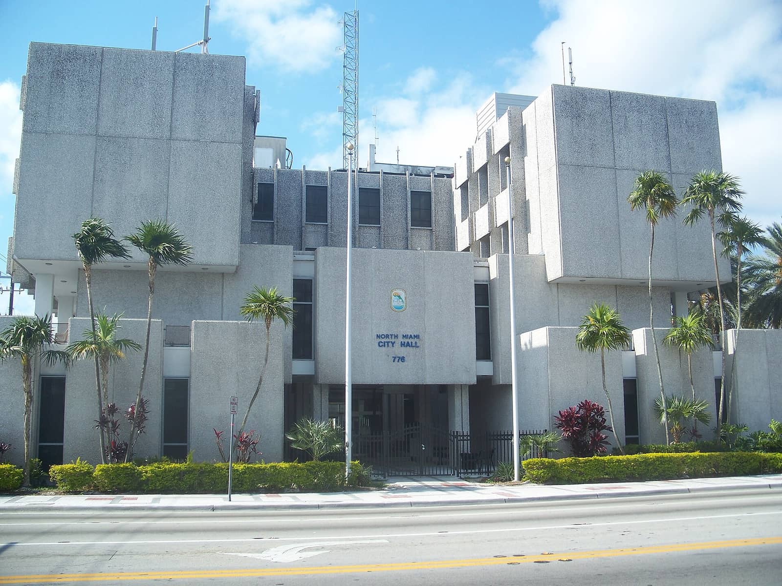 Concrete city hall building with a central entrance, palm trees, hedges, and a seal above the door.