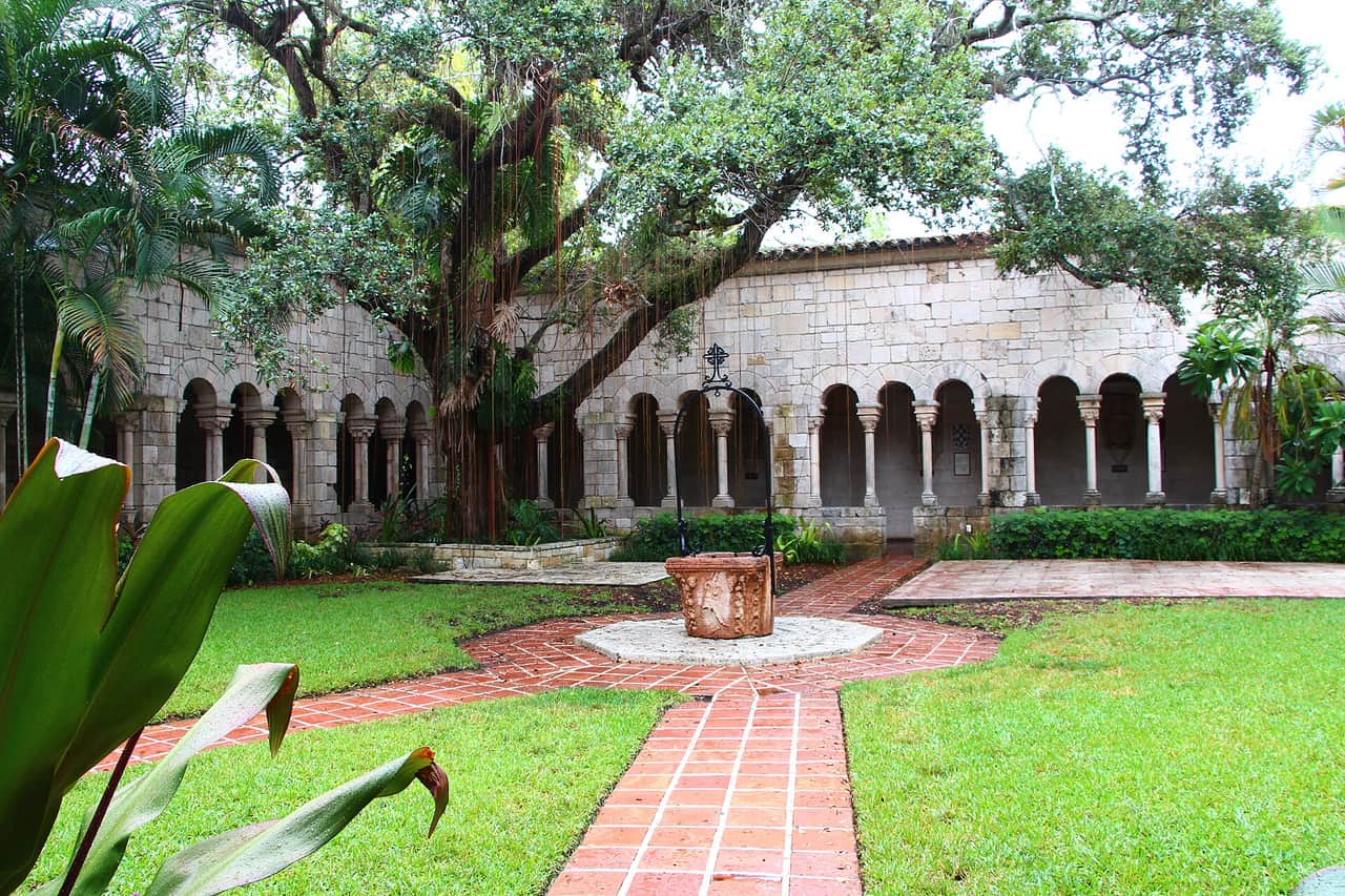 Stone cloister arcade around a courtyard with a large spreading tree and brick paths.