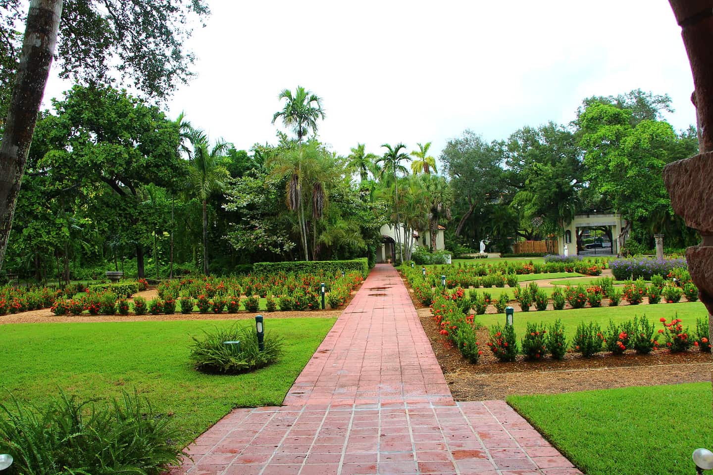 Formal garden with a brick pathway, green lawn, flower beds and palm trees under a cloudy sky.