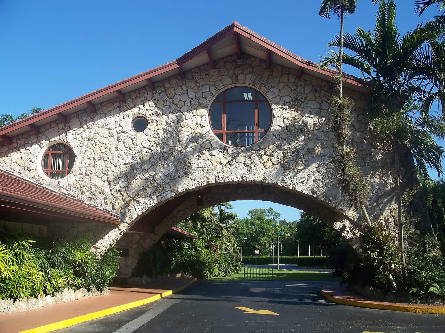 Stone archway gate with round windows and palm trees under a clear blue sky, leading to a road.