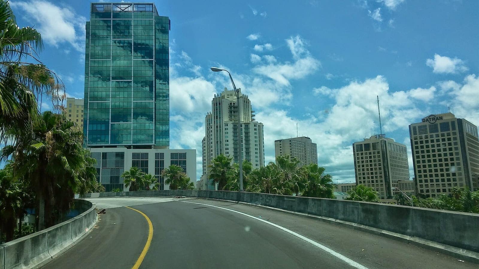 Highway ramp curves toward glass and stone high-rise towers under a blue sky with clouds and palms.