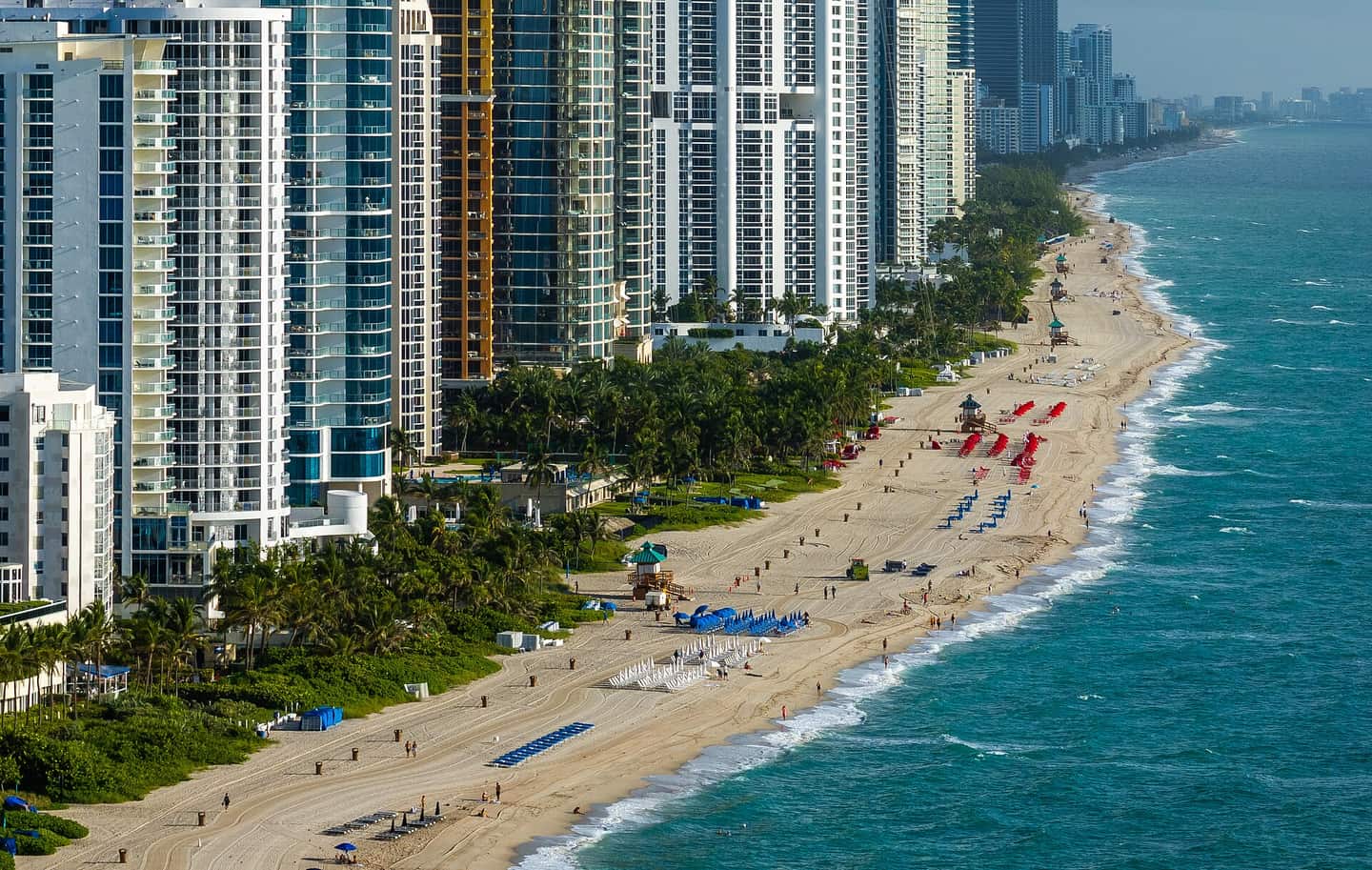 Aerial view of high-rise condos along a sandy beach with people and umbrellas.