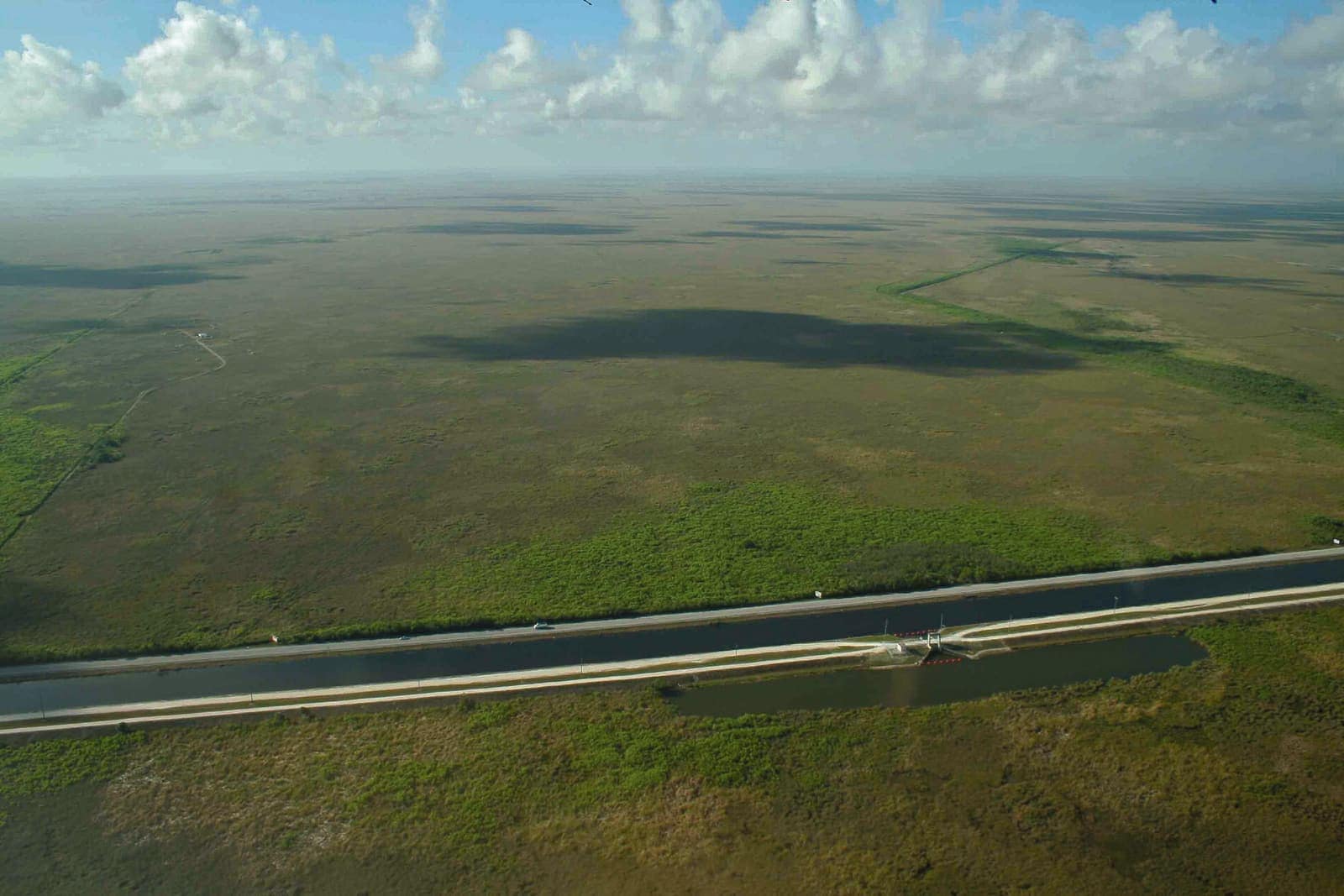 Aerial view of flat green wetland with a long straight road and canal under cloudy sky.