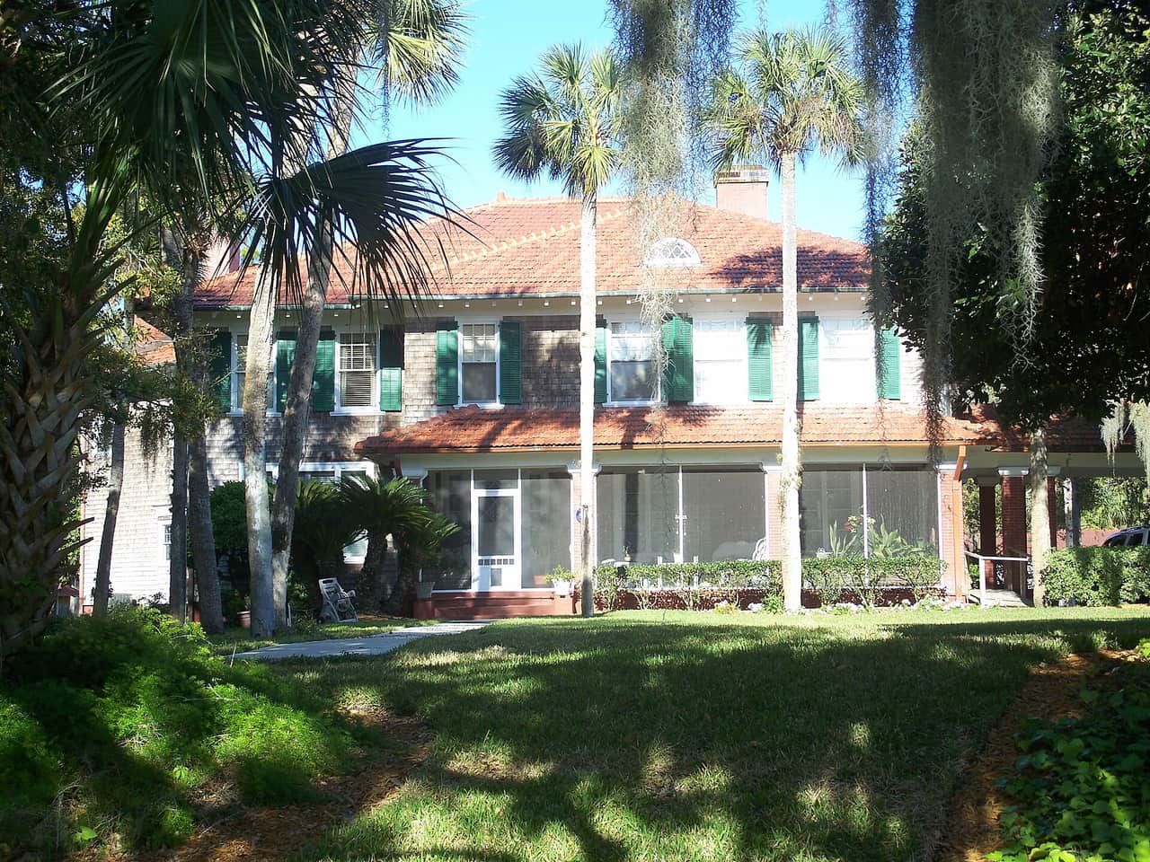 Two-story house with red tile roof, green shutters, screened porch, and palm trees on a sunny lawn.