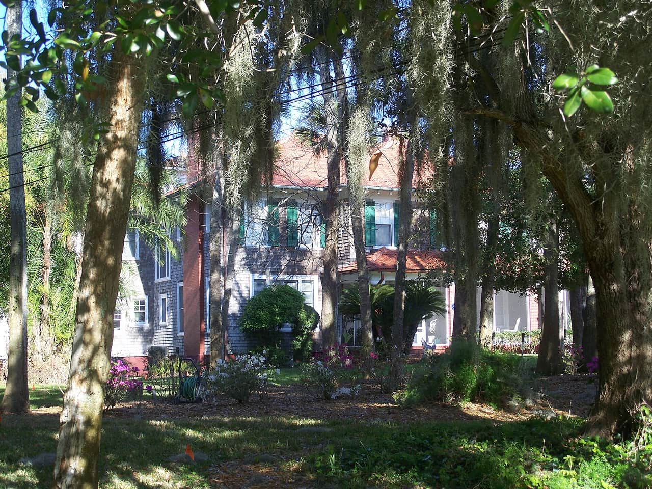 Two-story historic house with red tile roof and green shutters framed by live oaks and Spanish moss.
