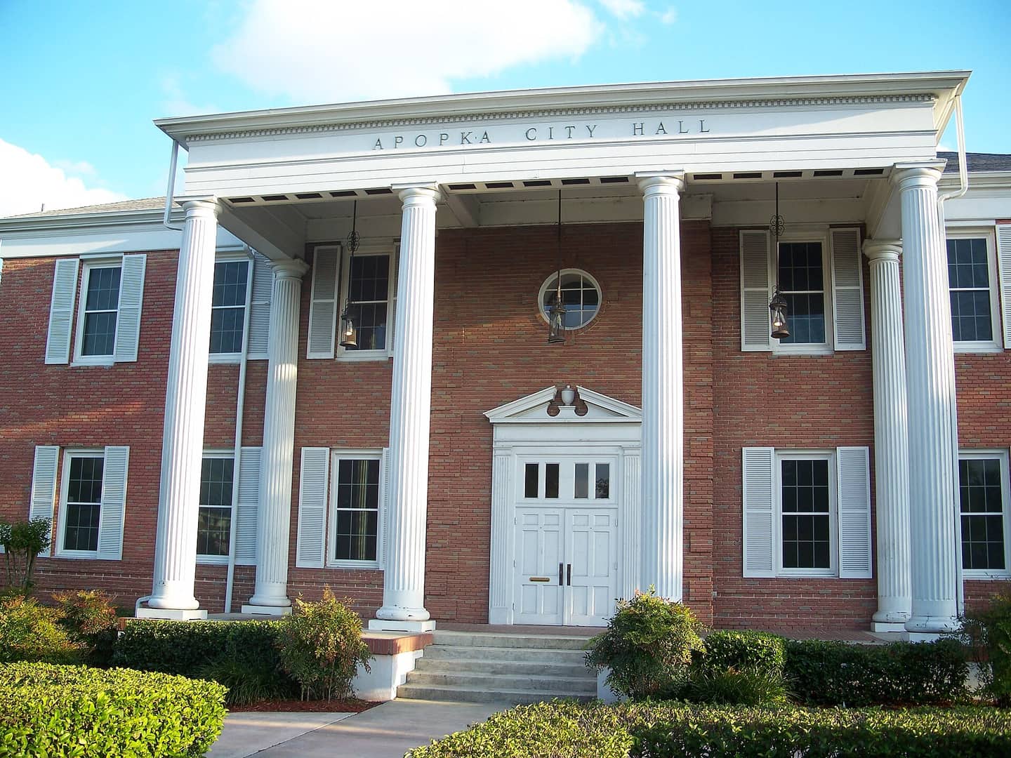 A two-story brick city hall with white columns and a pediment under a partly cloudy blue sky.
