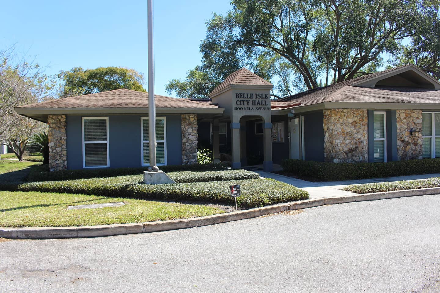 Single-storey City Hall building with stone columns, a flagpole, and a sign reading Belle Isle City Hall under clear blue sky.