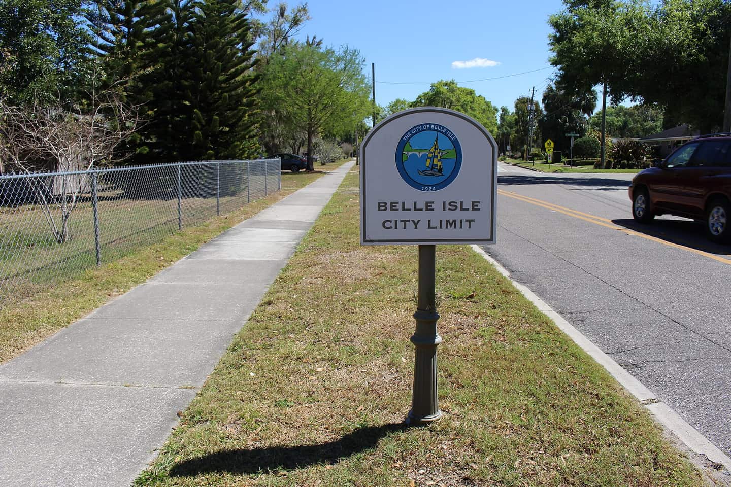 A roadside sign reading Belle Isle City Limit beside a sidewalk, with a red car on the right.
