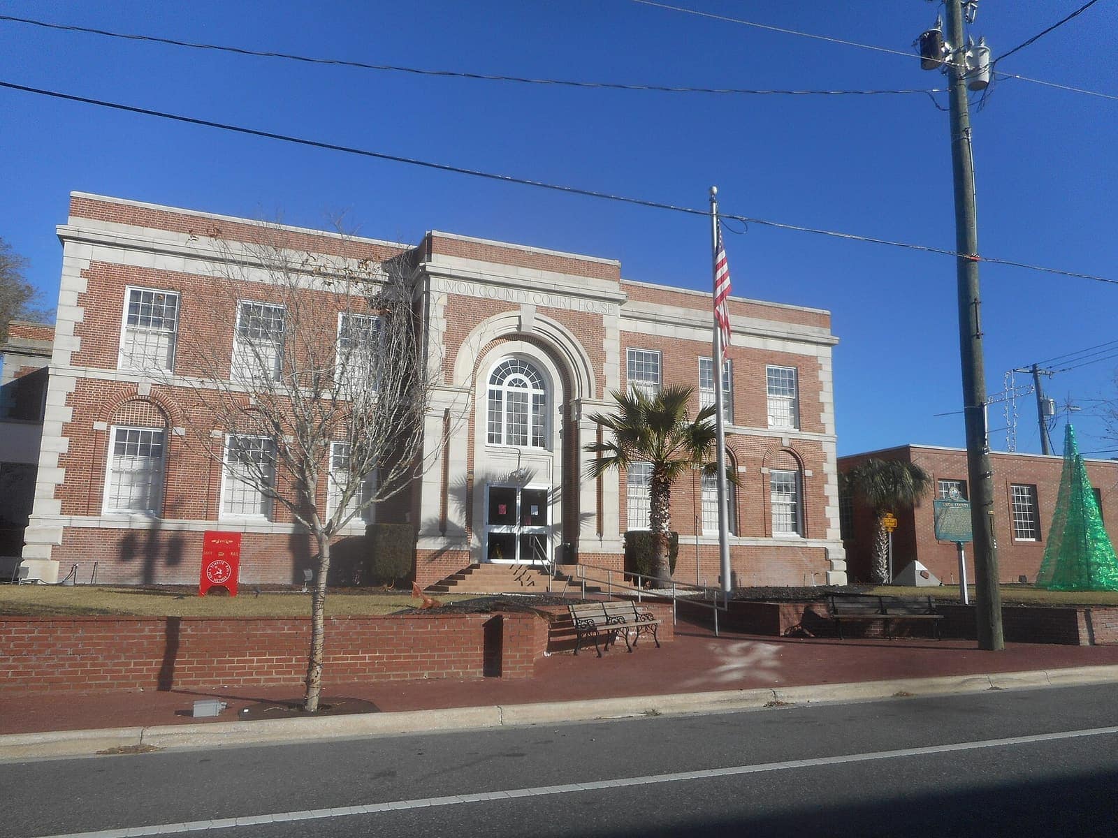 Brick courthouse with arched entrance, flagpole, benches and palm trees under blue sky.