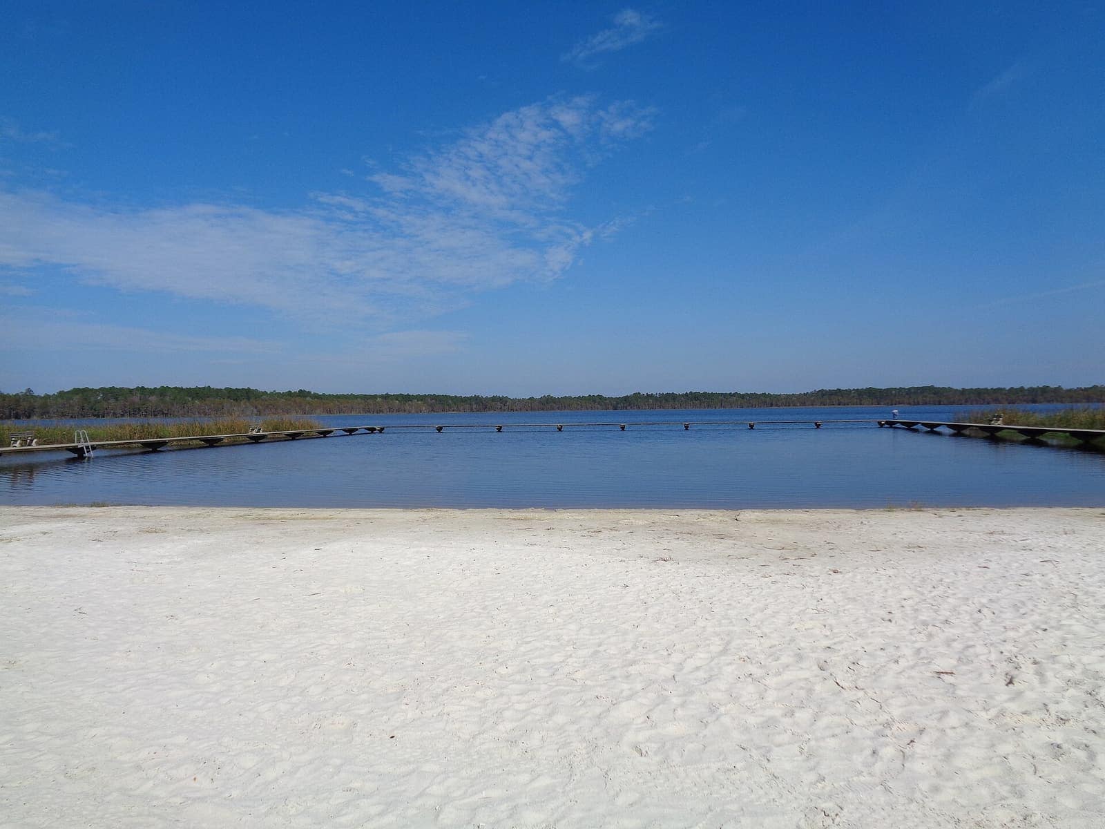 Pale sandy beach in front of a calm blue lake with distant docks and a wispy-cloud sky.