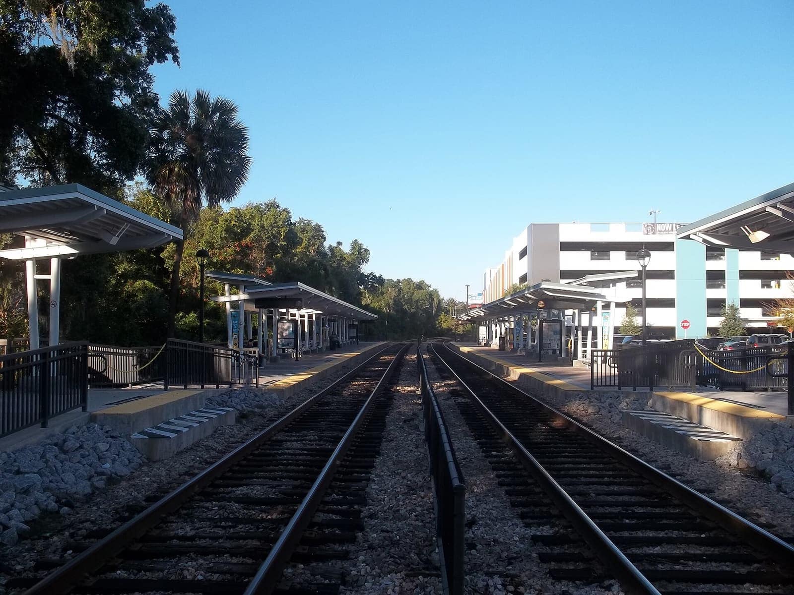 Two railway tracks run past sunlit platform canopies toward a white multi-story parking garage under a clear blue sky.