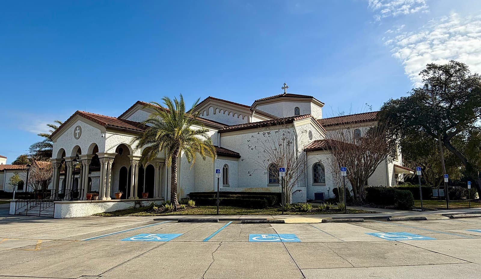 White stucco church with red tile roofs and arched windows beside an empty parking lot with blue accessible spaces.