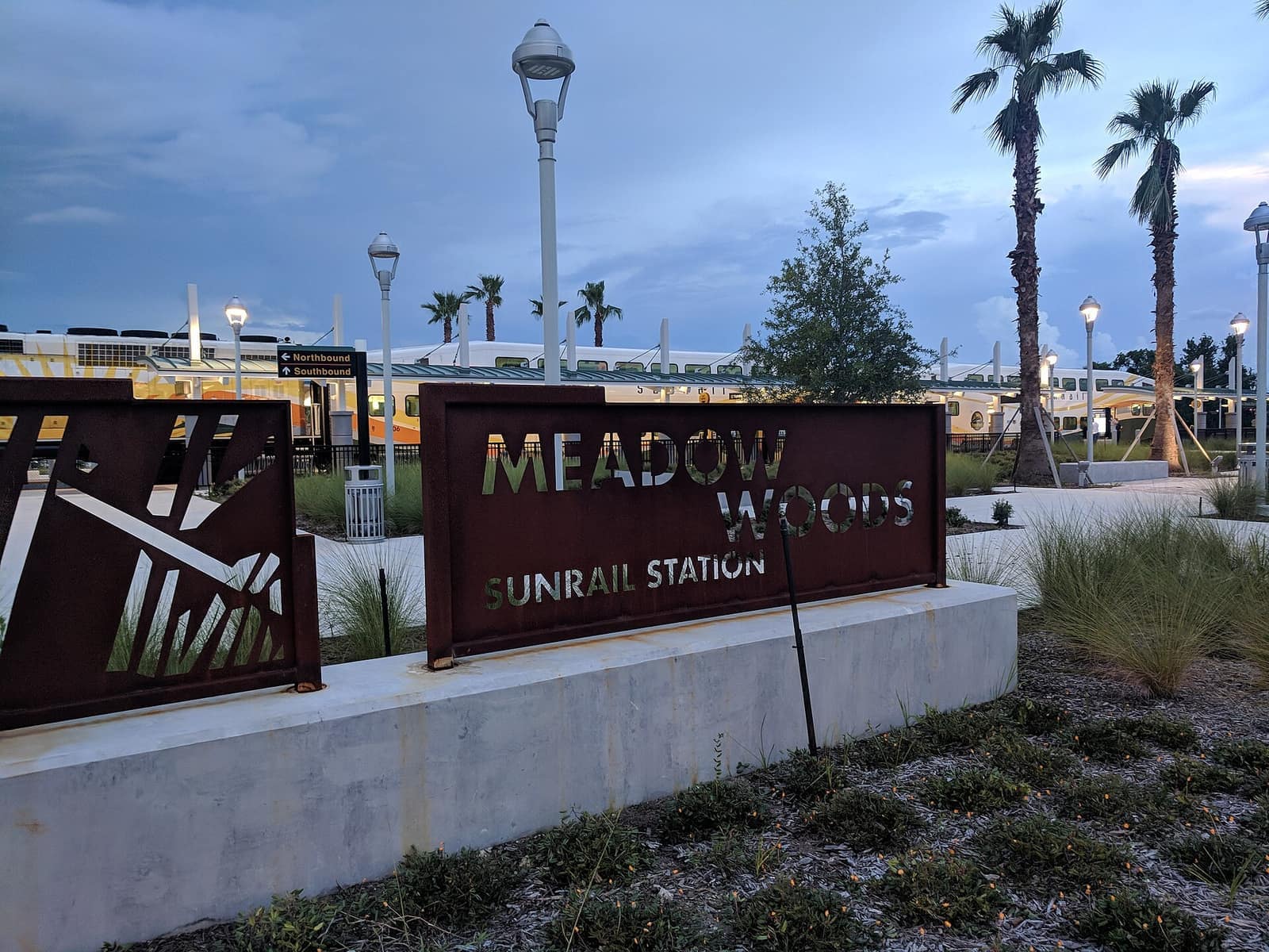 Metal Meadow Woods SunRail Station sign beside palm trees, with a passing train at dusk.