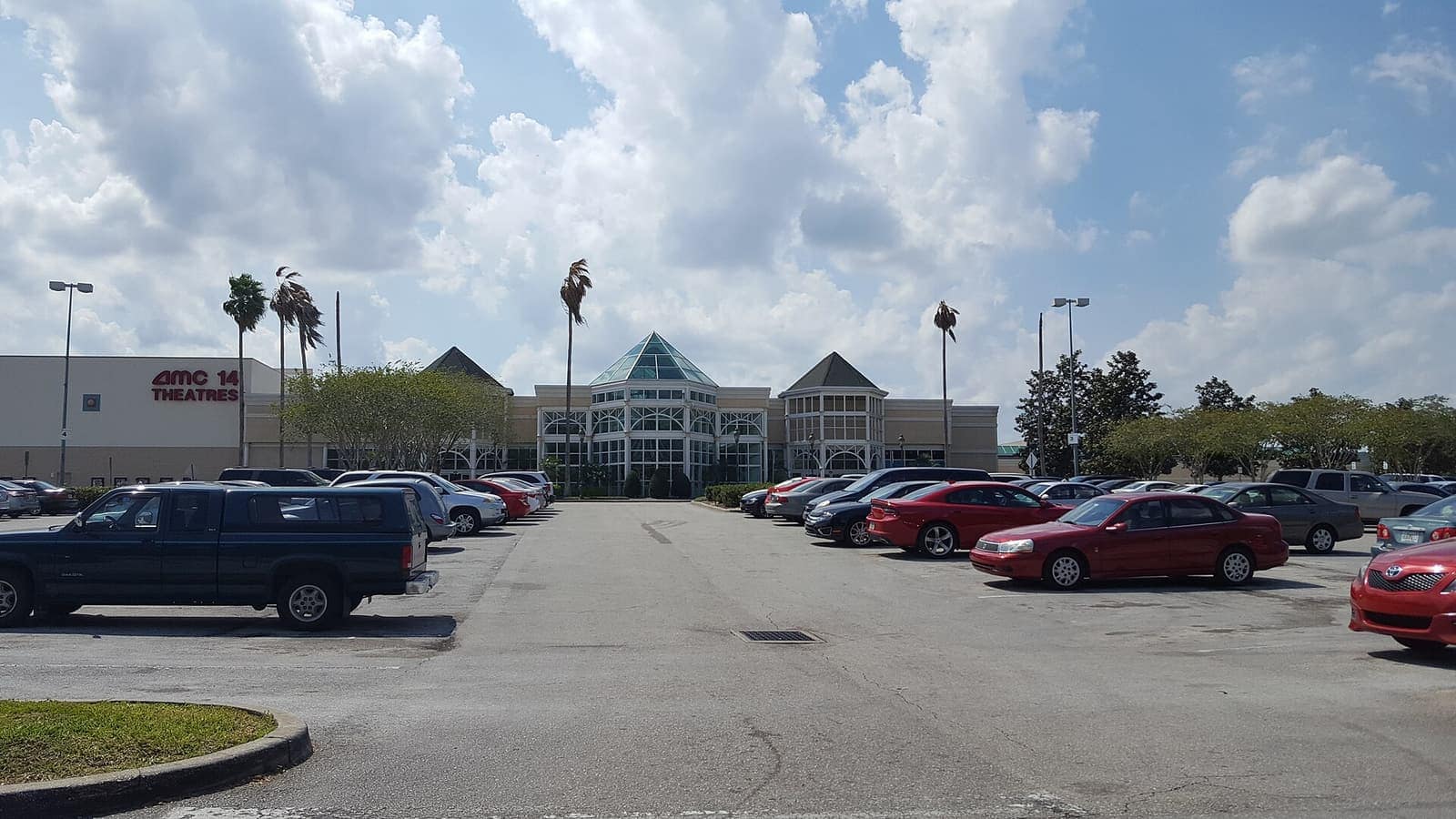 Large suburban shopping mall entrance with palm trees and a parking lot in front under cloudy sky.